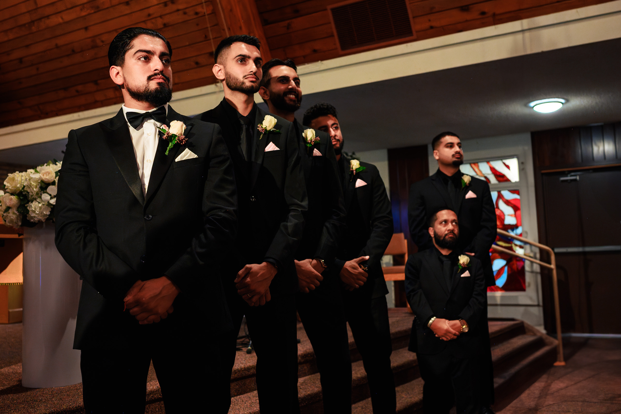 Groomsmen standing in a line inside a church, wearing black suits with boutonnières.