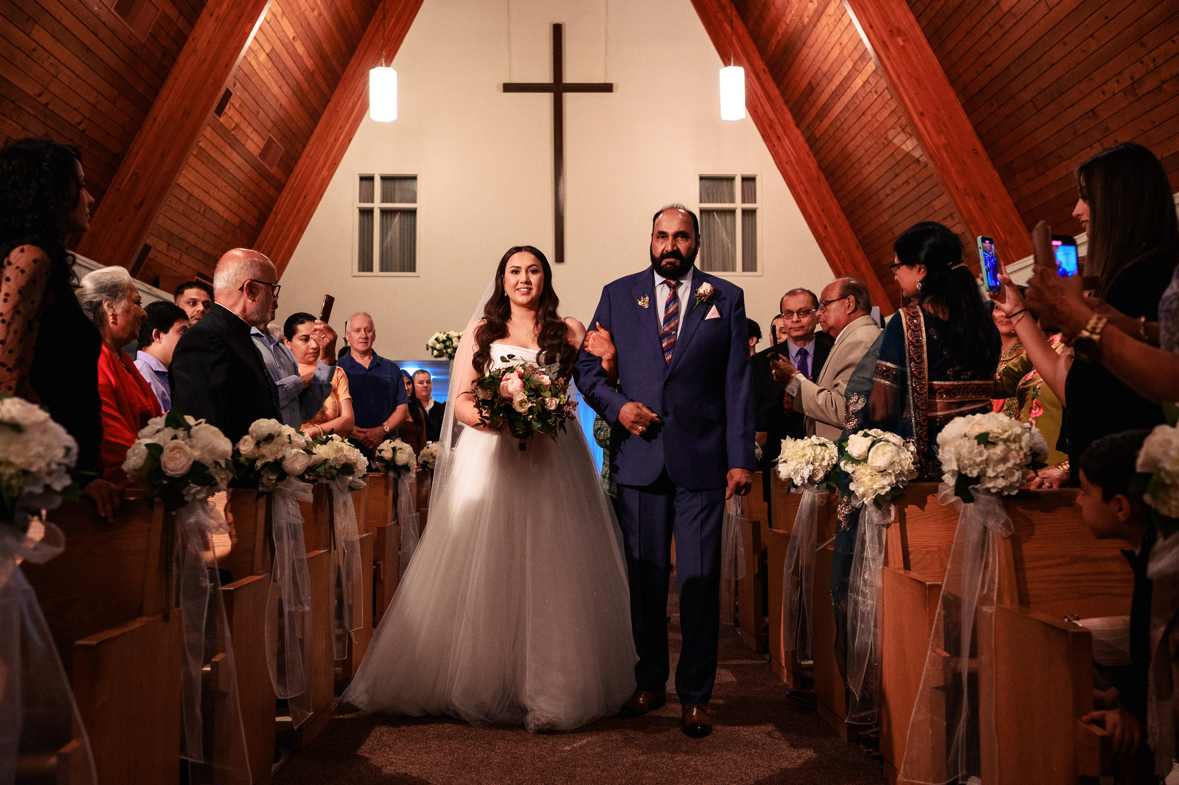 Bride and father walk down the aisle in a church with guests seated on either side.