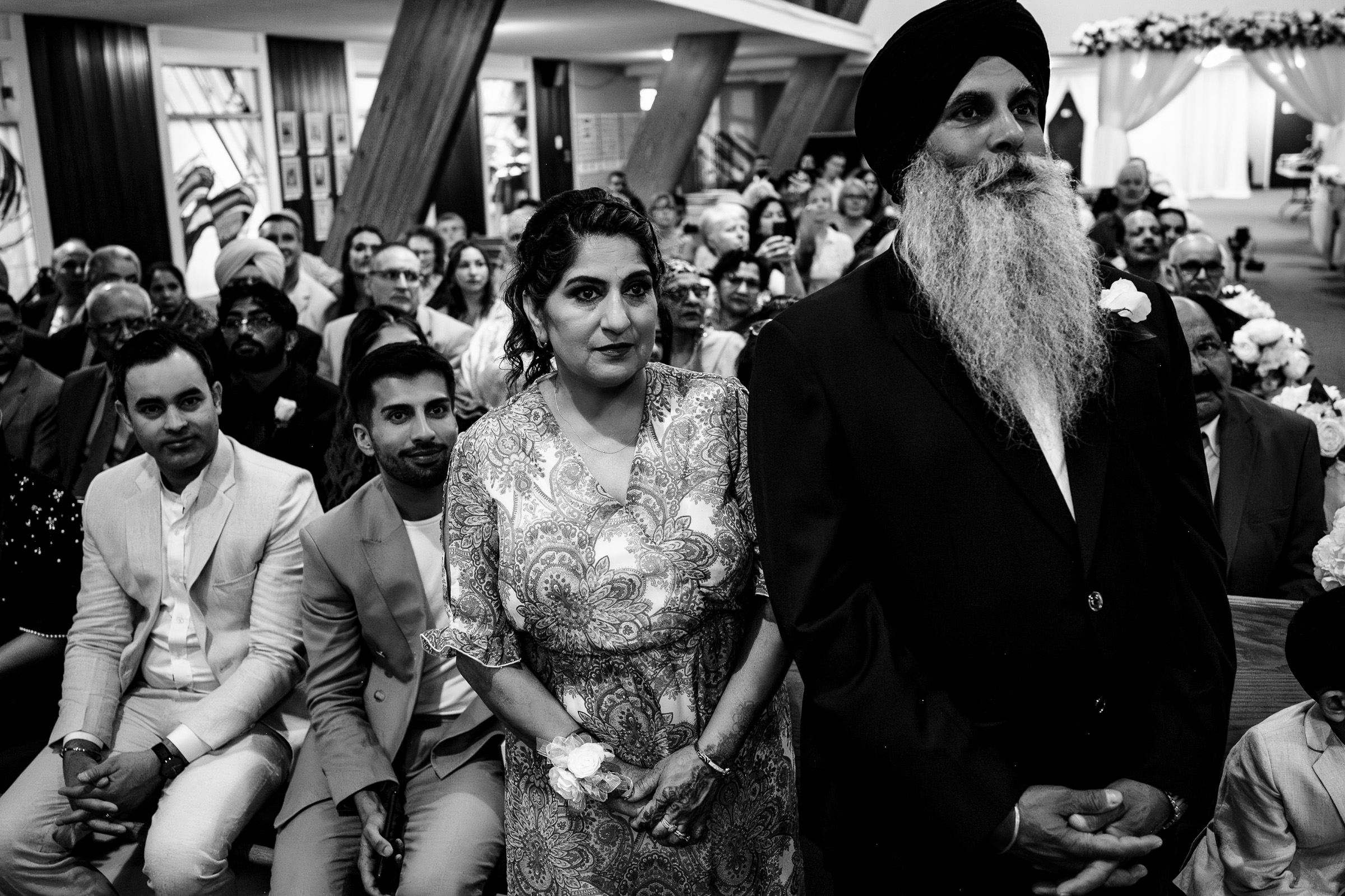 People seated in a hall, facing forward, with a man and woman standing in the foreground.