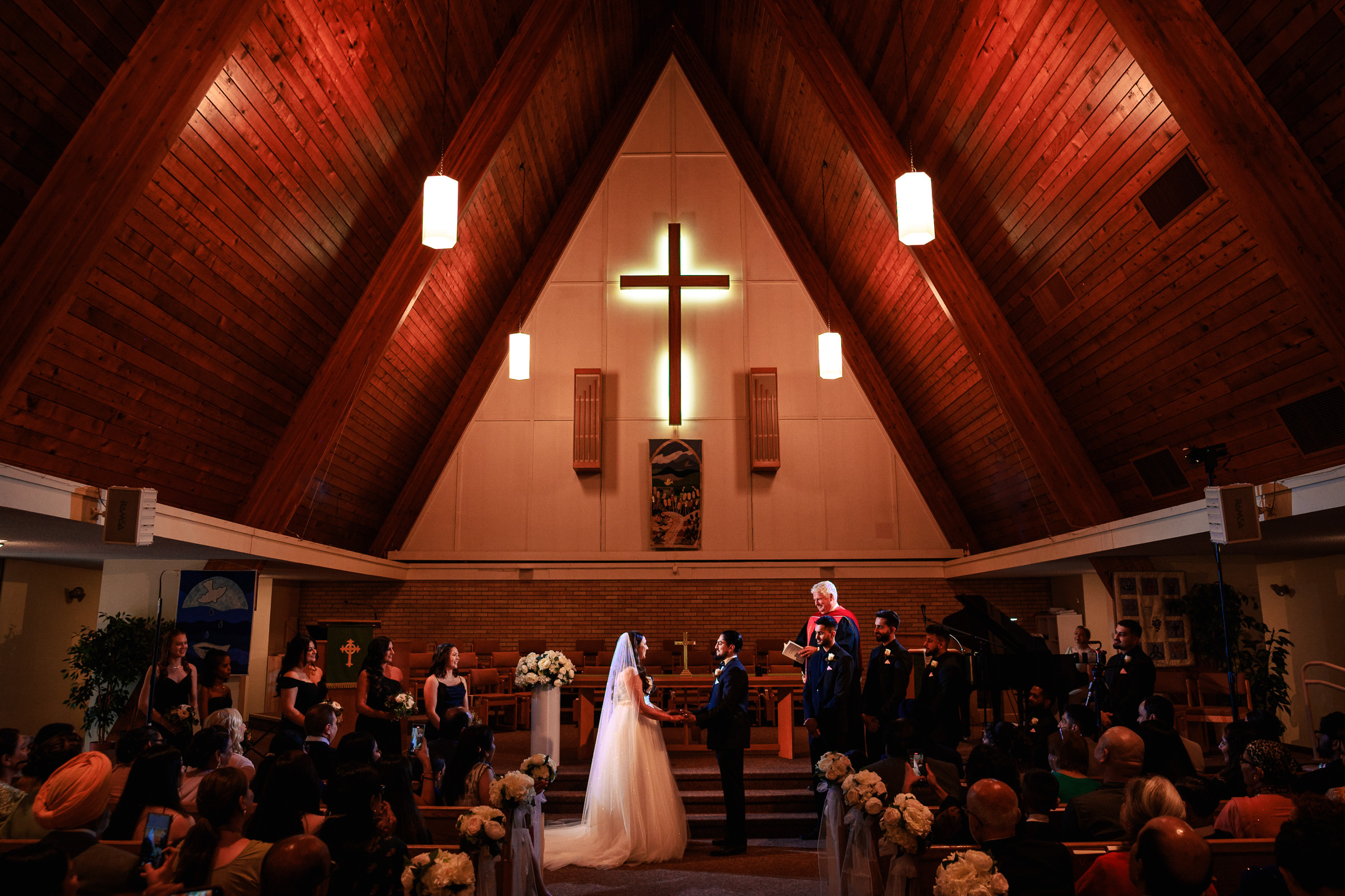 A couple stands at the altar in a wooden church with a glowing cross above them.