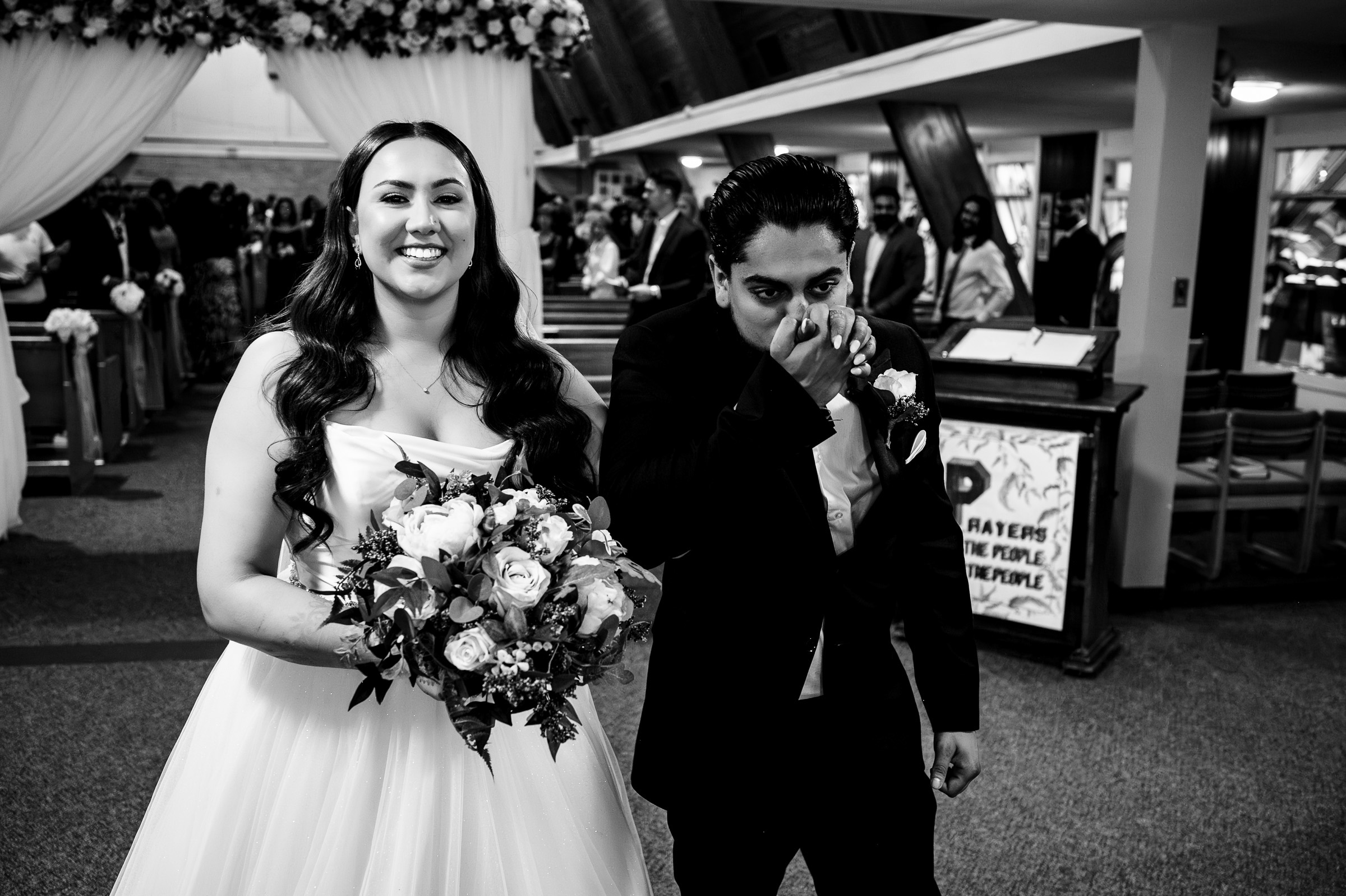 Bride and groom walking down the aisle, smiling and holding a bouquet, guests in the background.