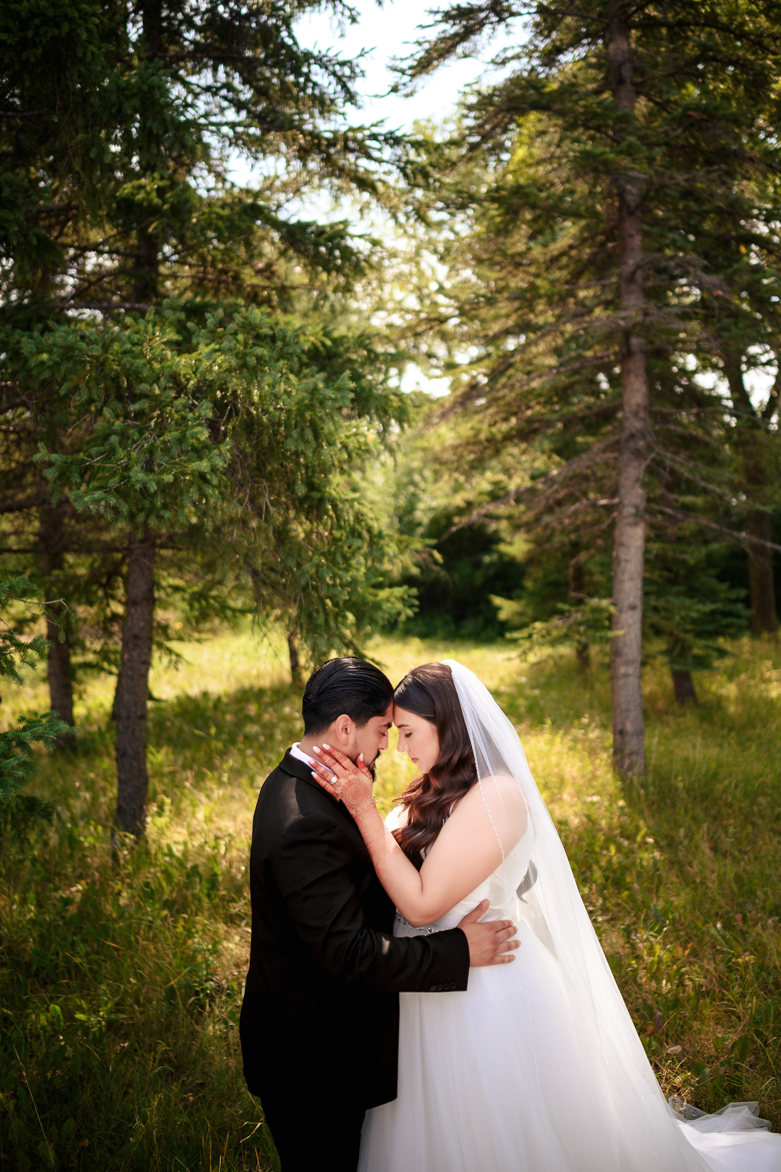 Bride and groom embrace in a forest, surrounded by tall trees and grass.