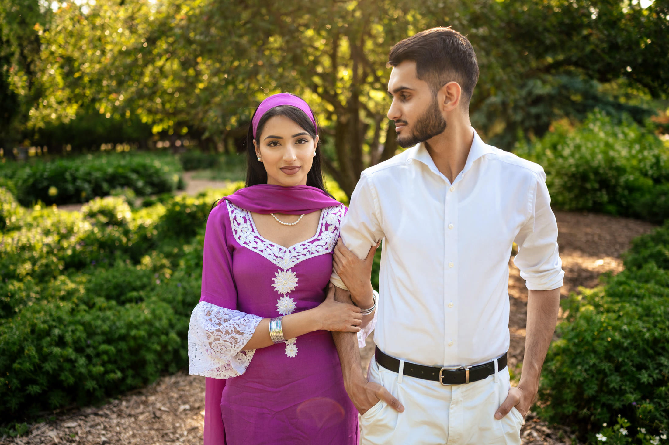 A couple strolls arm-in-arm in a sunlit garden, celebrating their engagement at home.