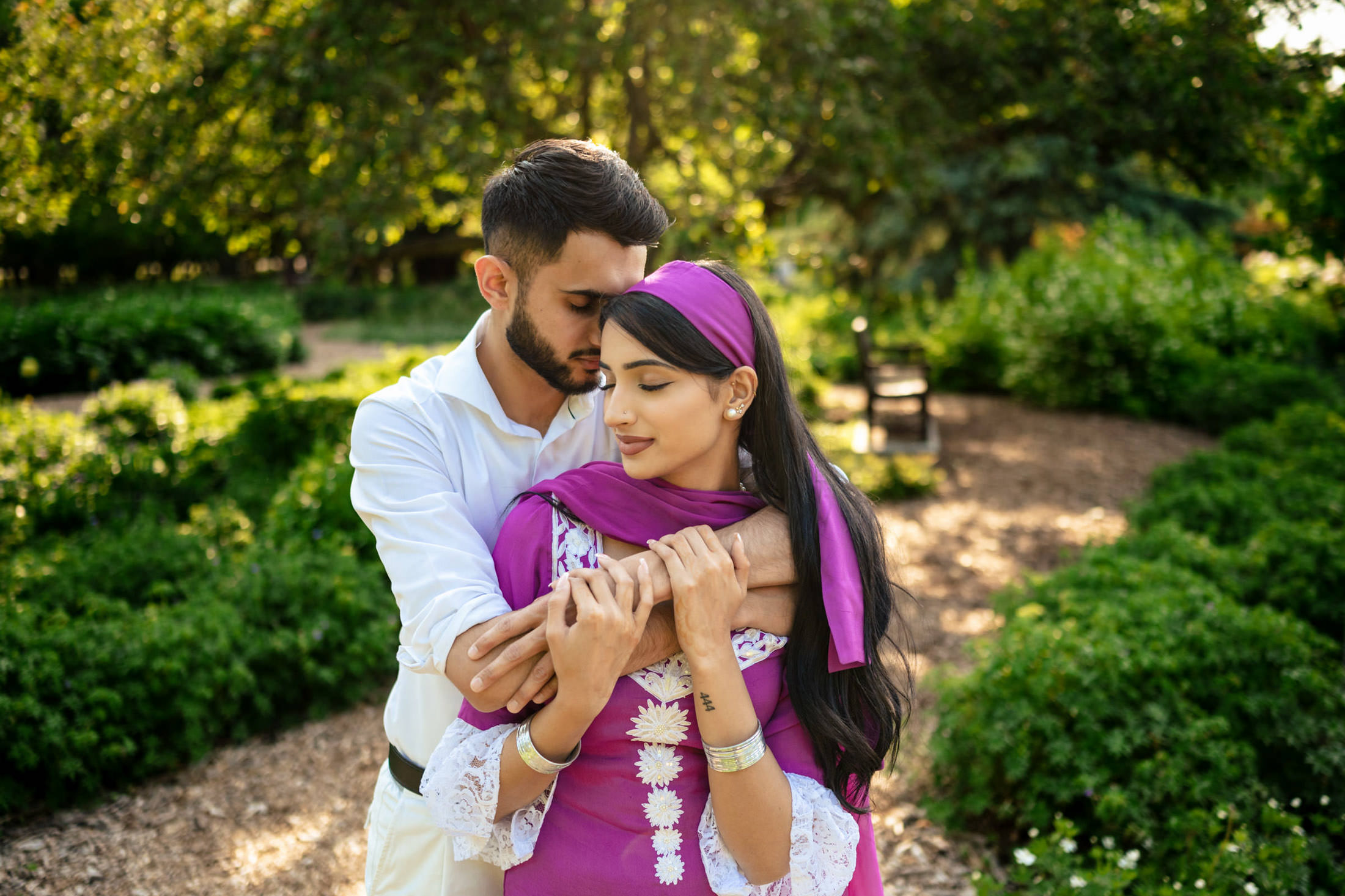 A couple embraces in a lush green garden during their engagement under soft sunlight.
