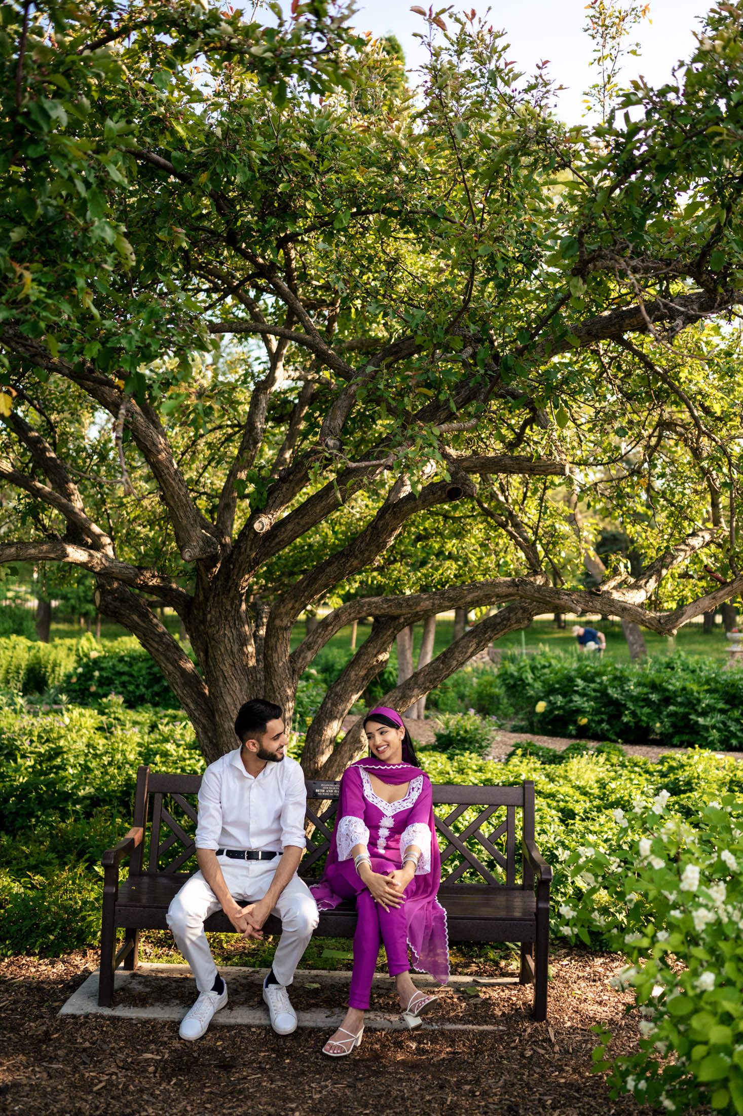 A couple in bright attire sits on a bench under a large tree, celebrating their engagement.