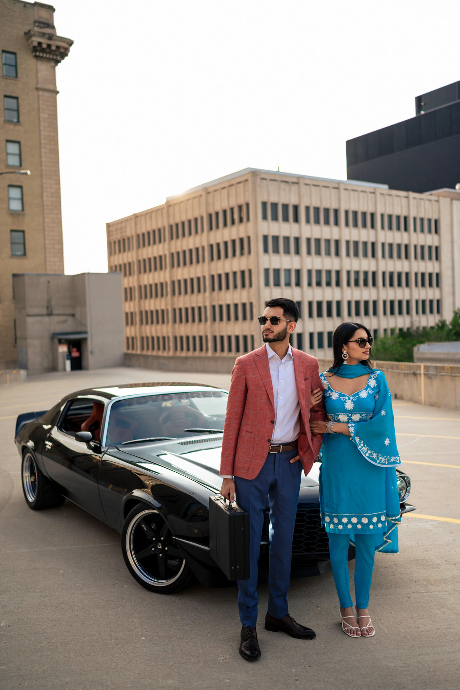 A couple in formal attire stands by a vintage black car after an engagement at home.