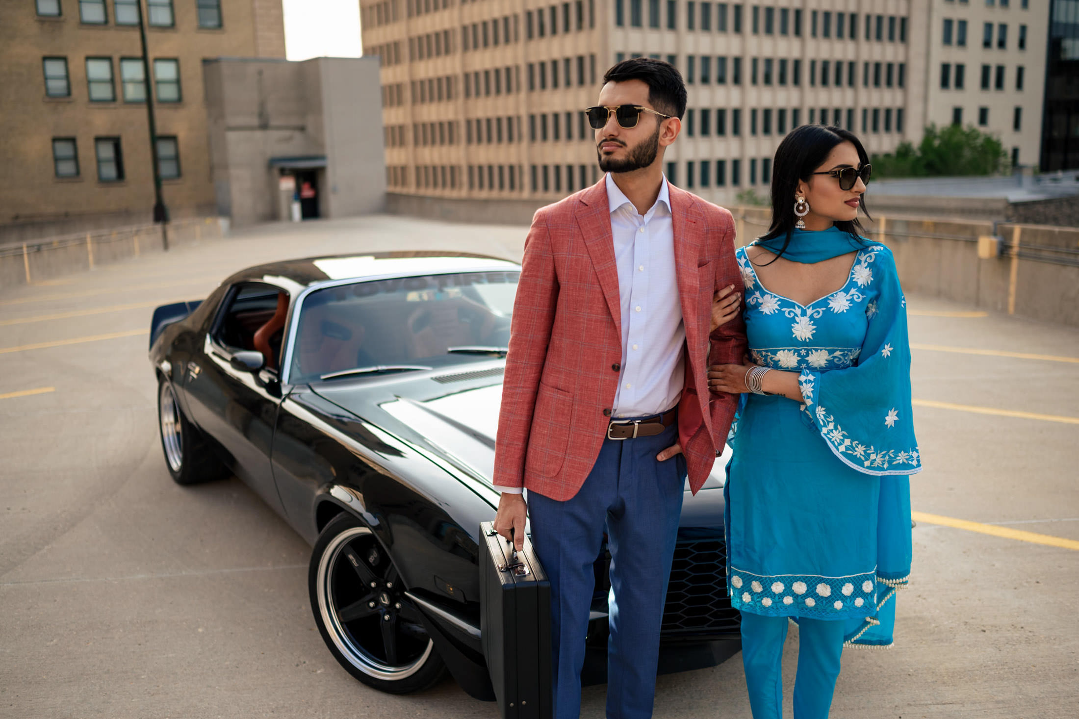 A couple in stylish attire celebrates their engagement by a sleek black car on a rooftop.