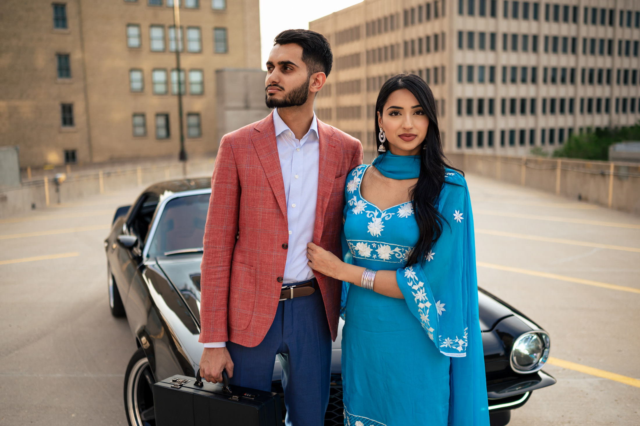 Couple poses stylishly by a black car, capturing an at-home engagement vibe; man in red jacket.