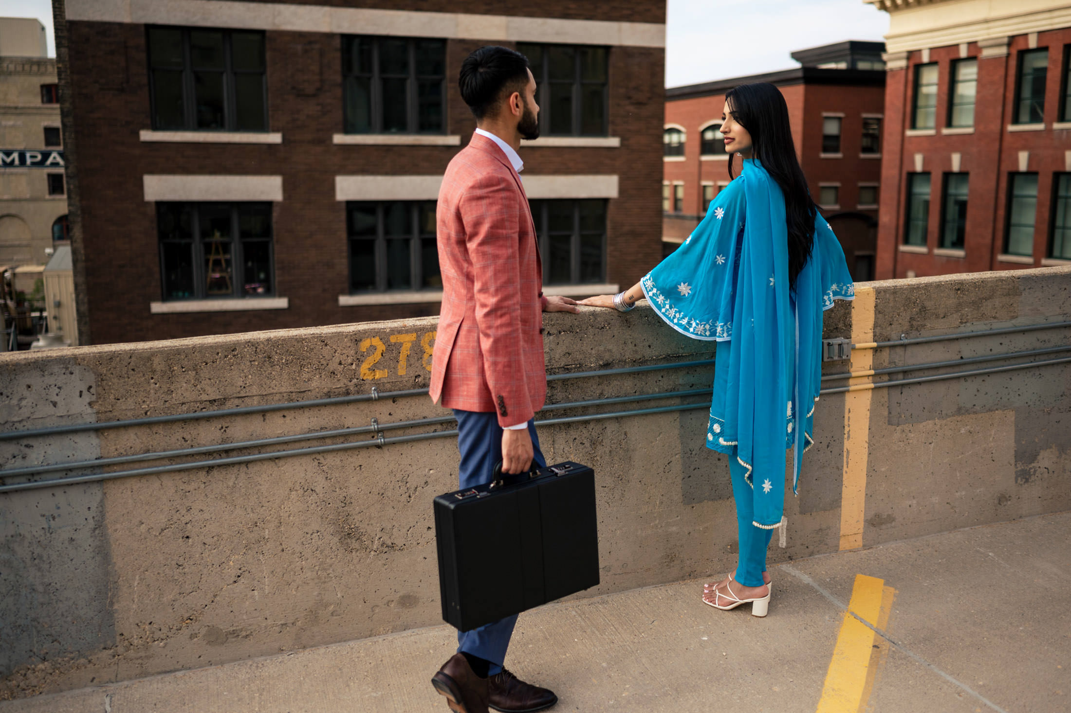 Man in pink blazer with briefcase chats with woman in blue attire on rooftop, enjoying engagement.