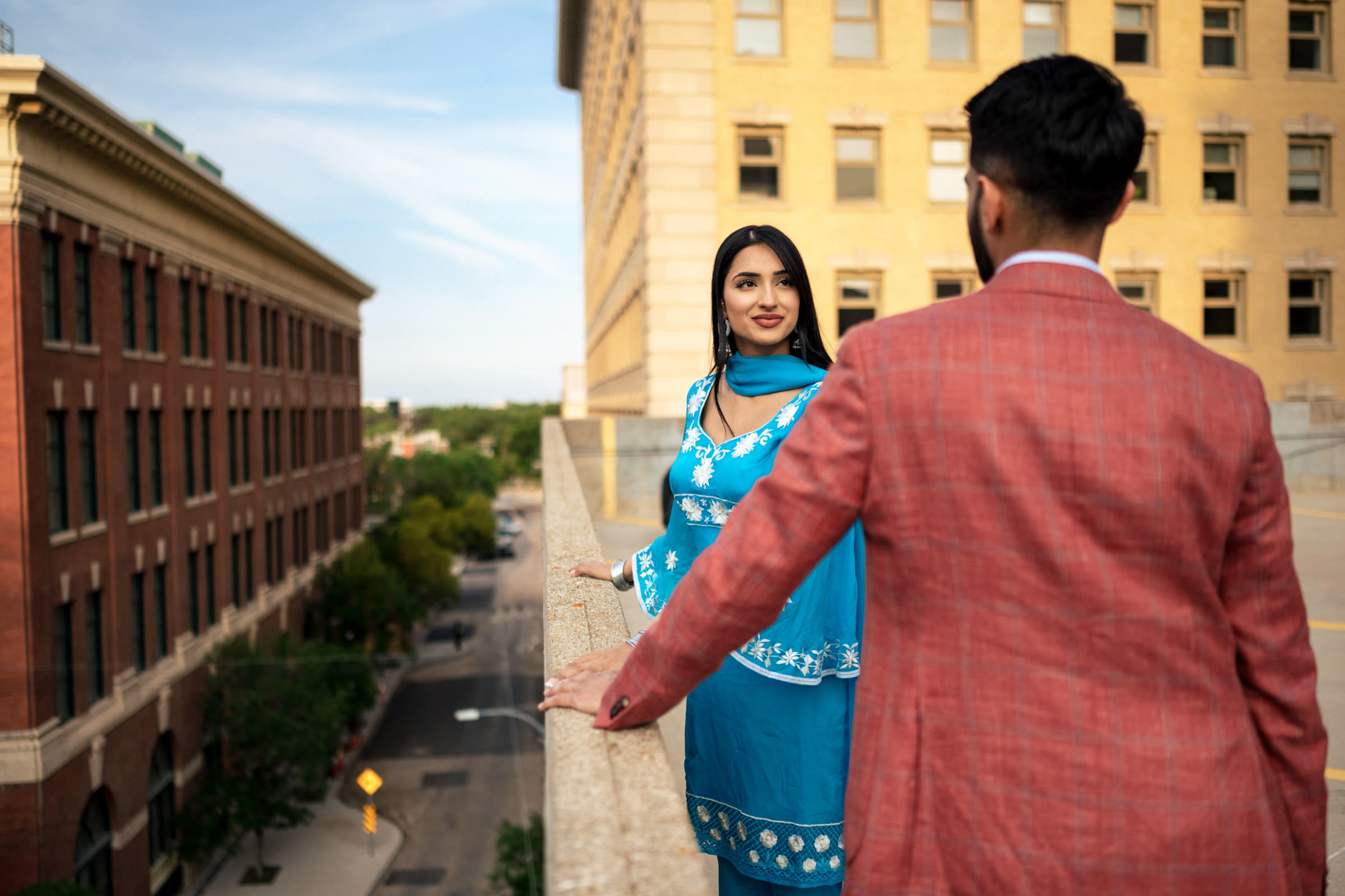 A woman in blue and a man in red share a rooftop moment, hinting at engagement.