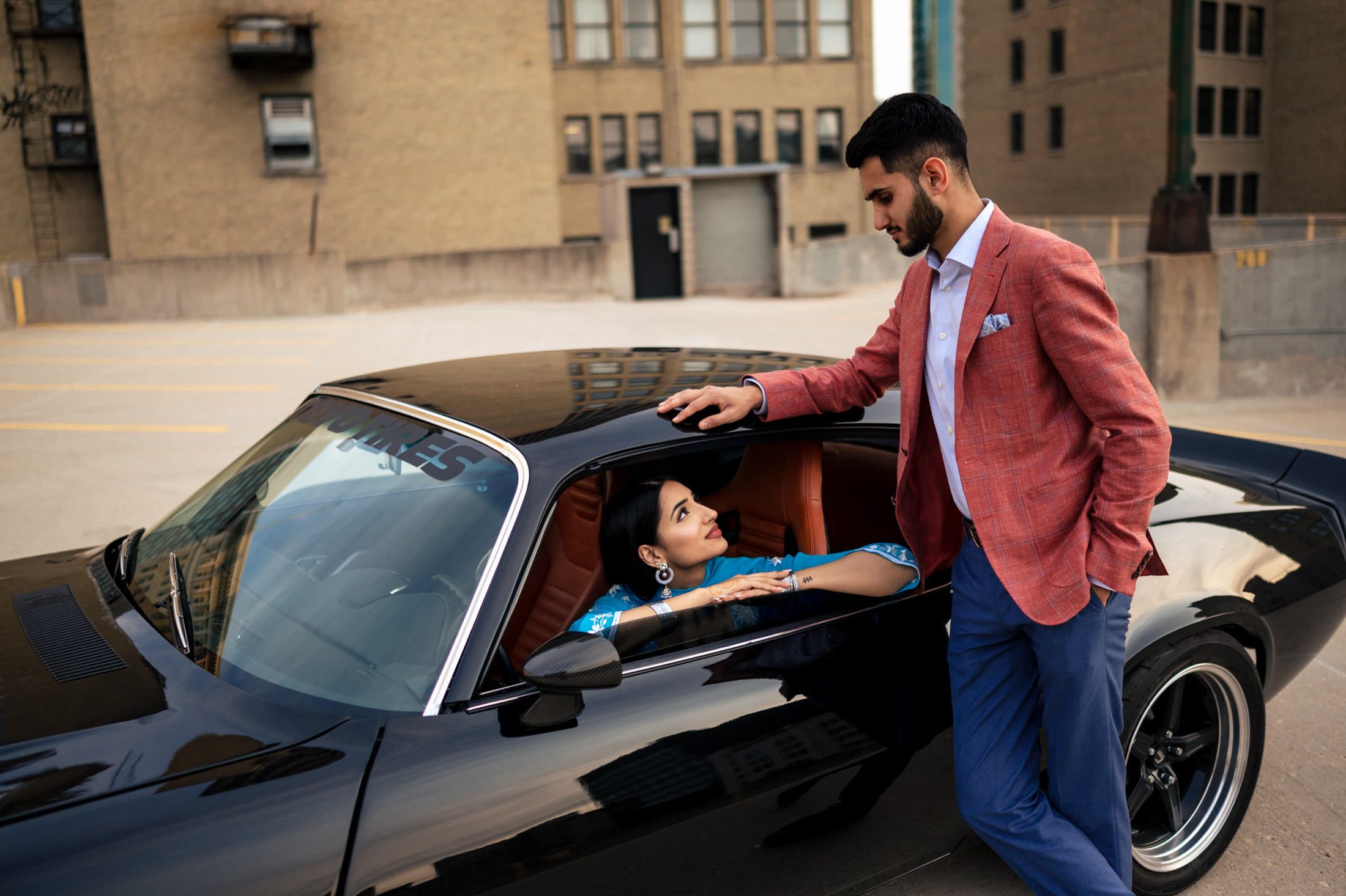 Man in a red jacket leans on a black car, discussing home engagement with woman inside.