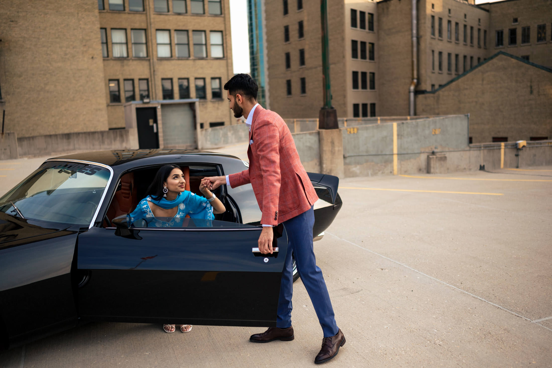 A man aids a woman in a blue dress exiting a black car, hinting at engagement at home.