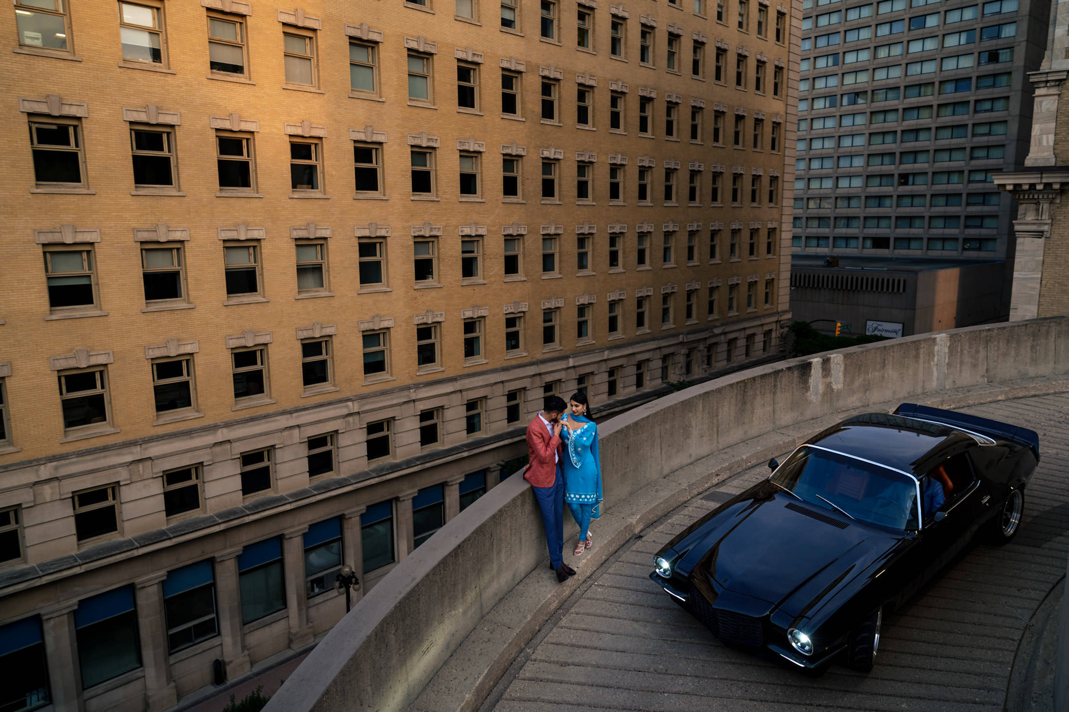 Couple marks engagement on curved road by classic car, with city backdrop.