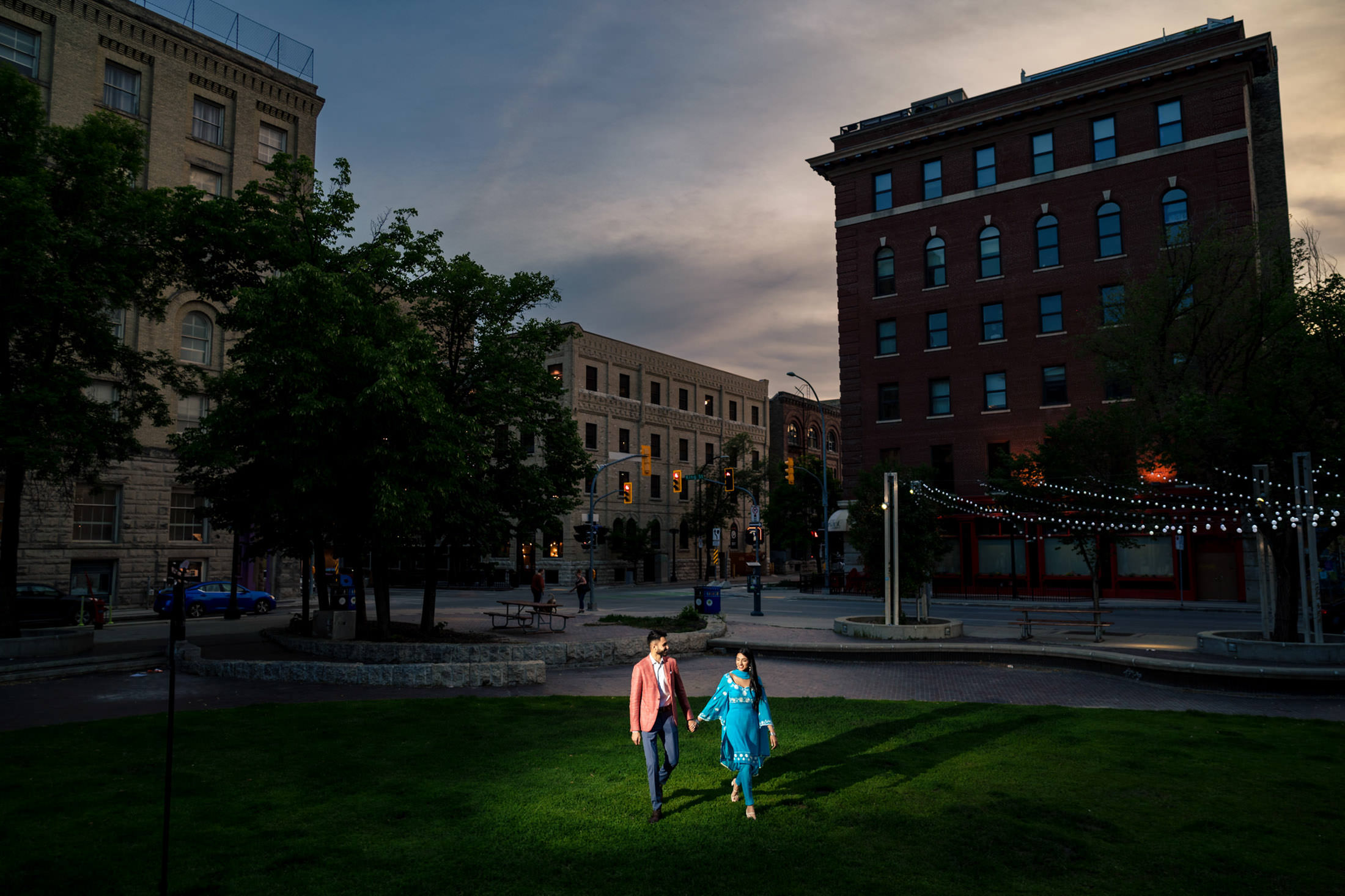 Engagement Photos at home: An engaged couple strolls hand in hand on a grassy area in the city at dusk.