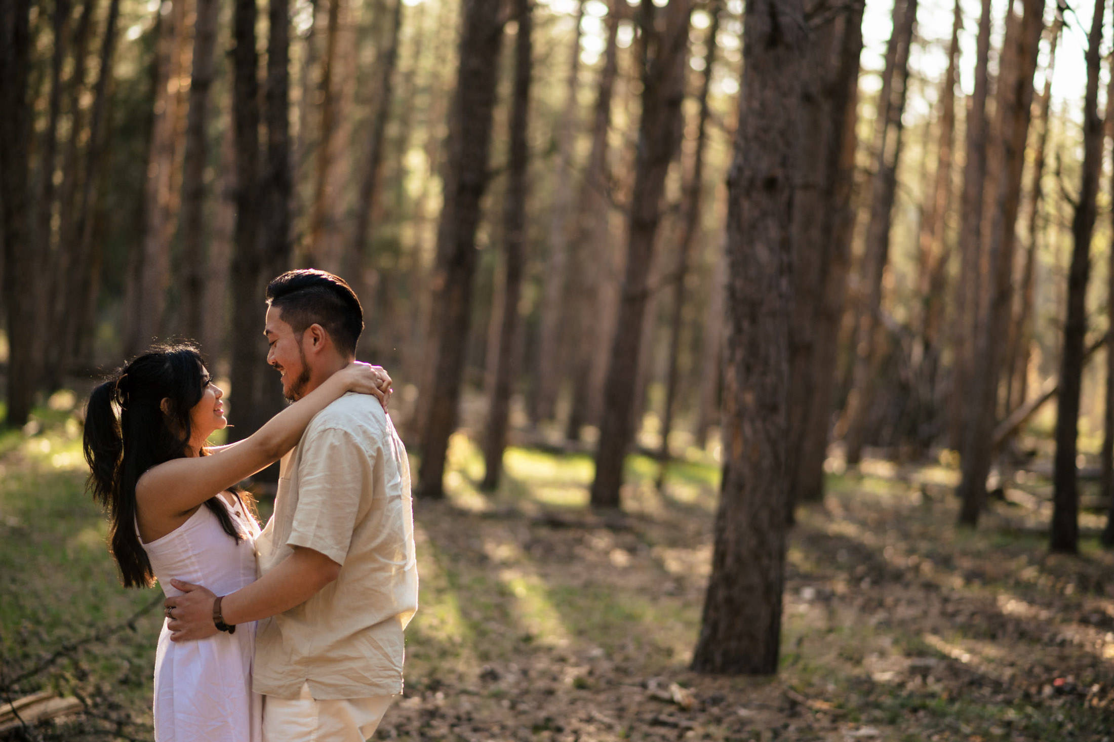 A couple embraces in a sunlit forest clearing, their dog playfully joining them.