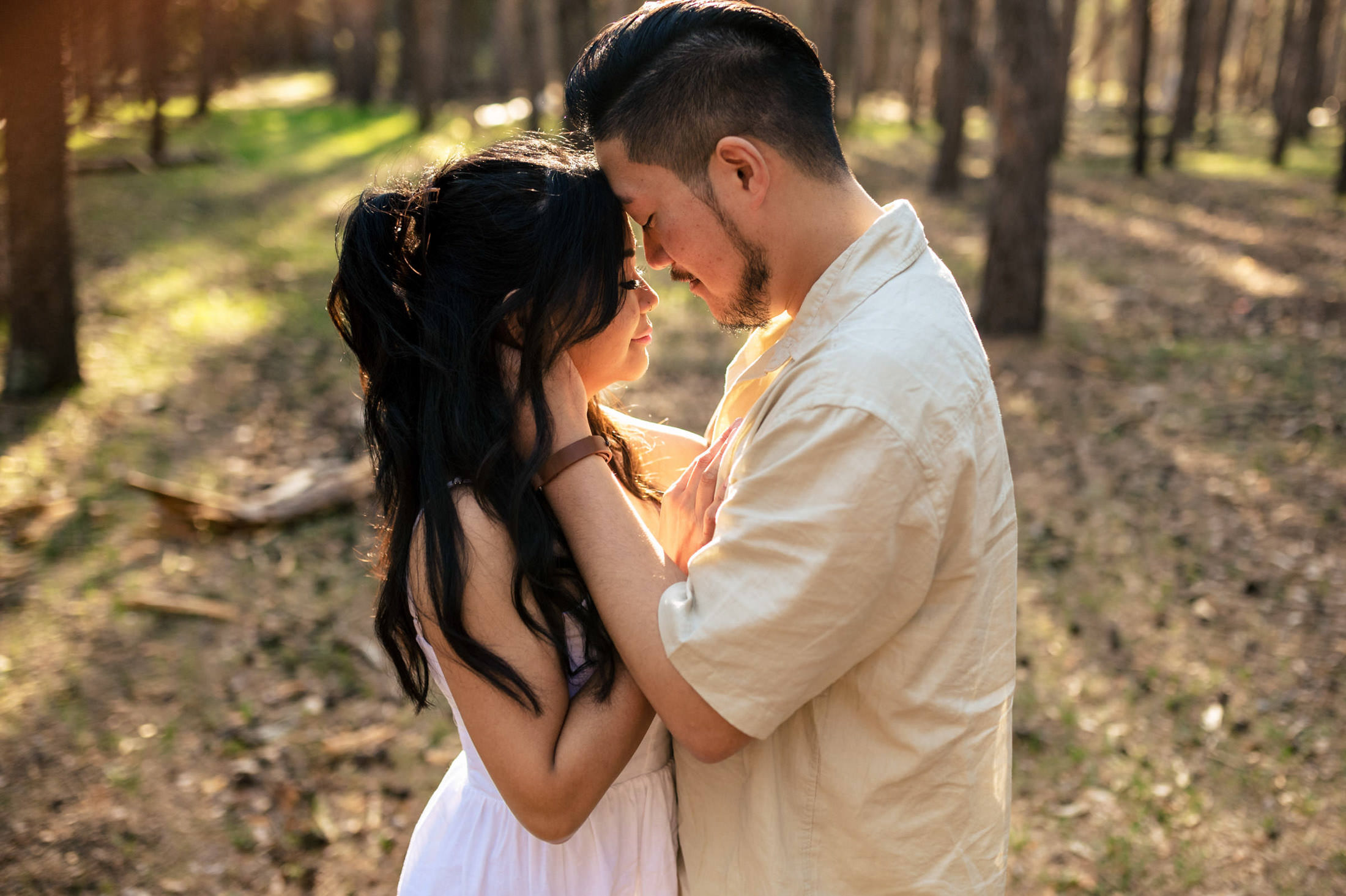 Couple embracing in a sunlit forest with their dog, eyes closed as sunlight filters through trees.