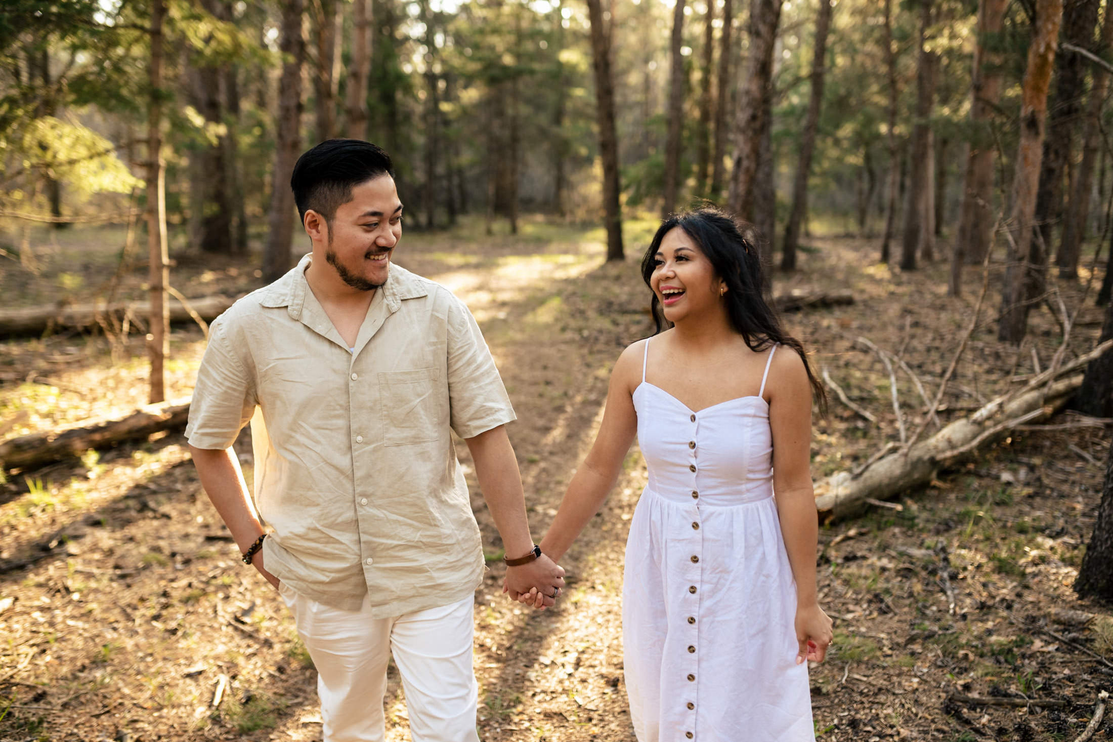 A couple engaged, smiling, and walking hand-in-hand with their dog in a sunlit forest.