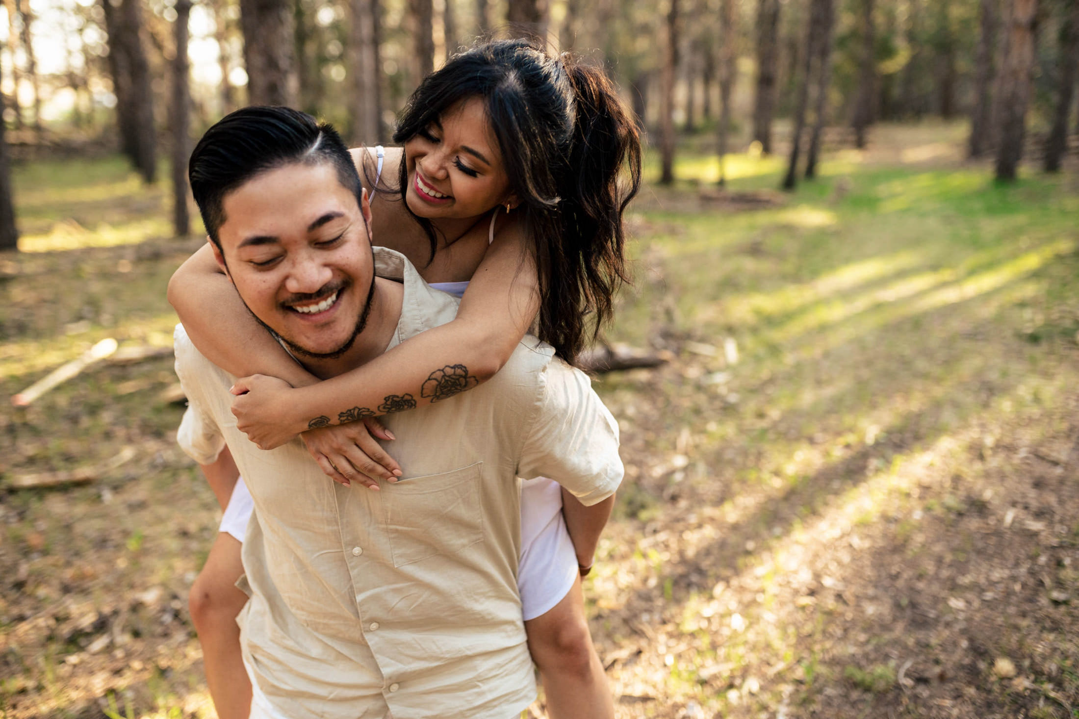 A smiling couple enjoys a playful piggyback ride with their dog in a sunlit forest.