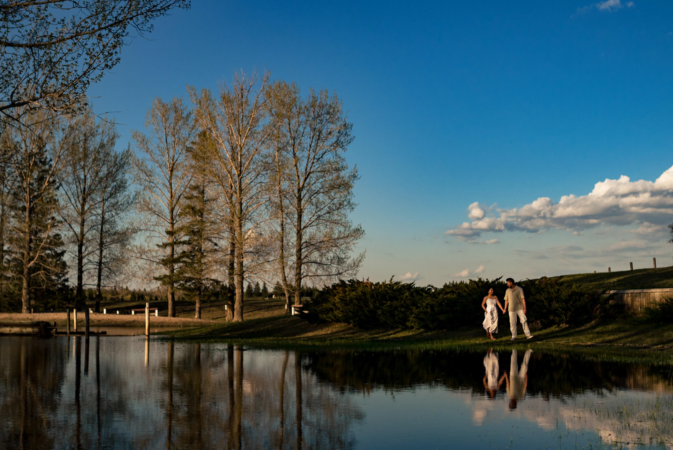 A couple with their dog walking by a pond under a clear blue sky.