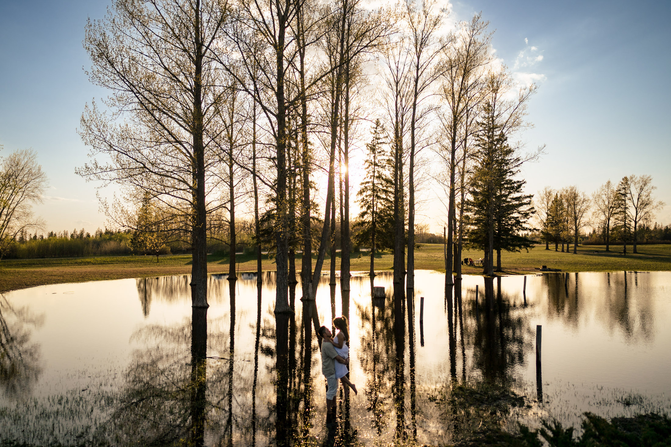 Child and dog stand among trees in a flooded park at sunset, with reflections on the water.