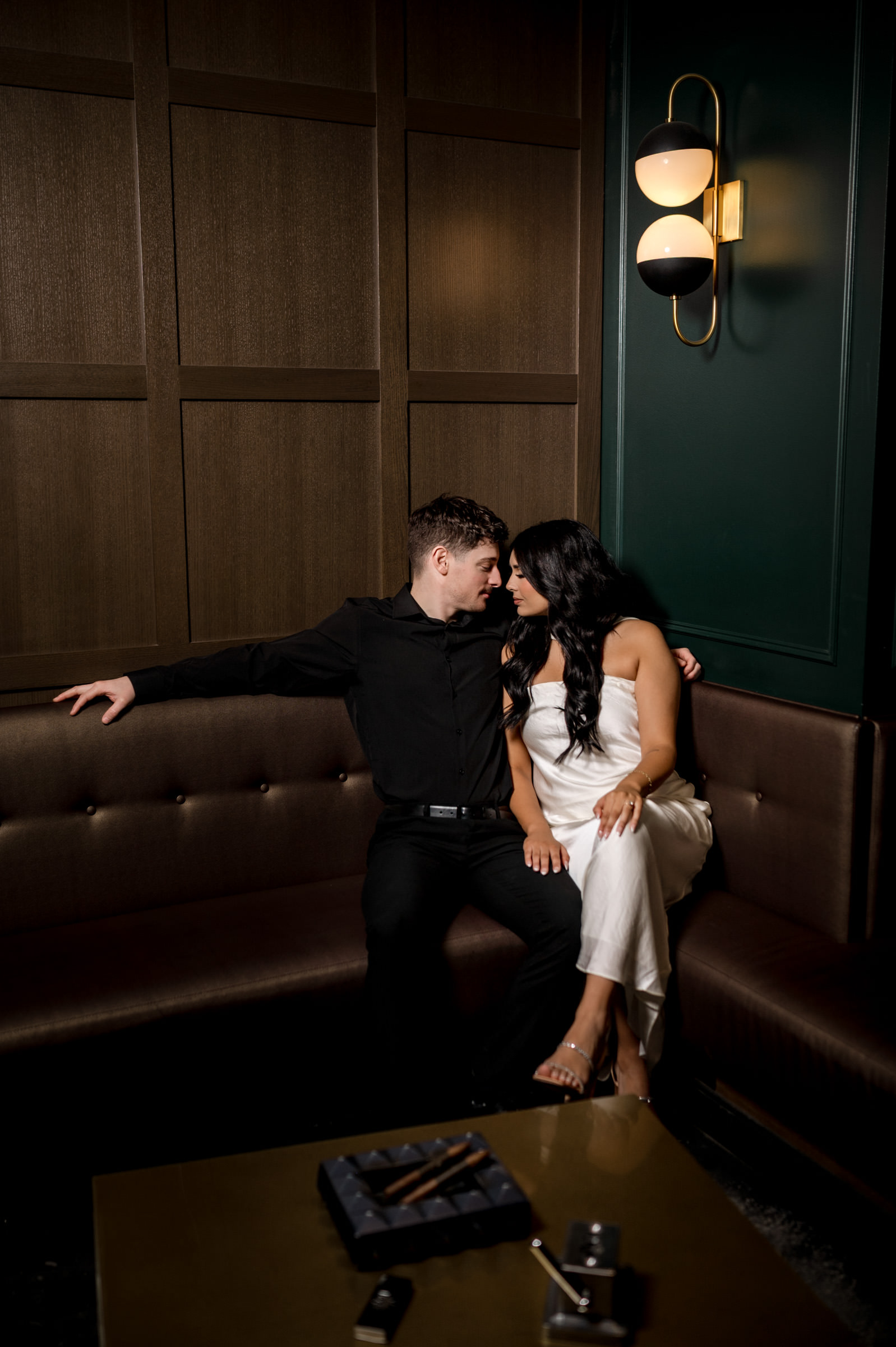 Couple sitting closely on a couch in a dimly lit room, sharing a Winnipeg engagement moment.