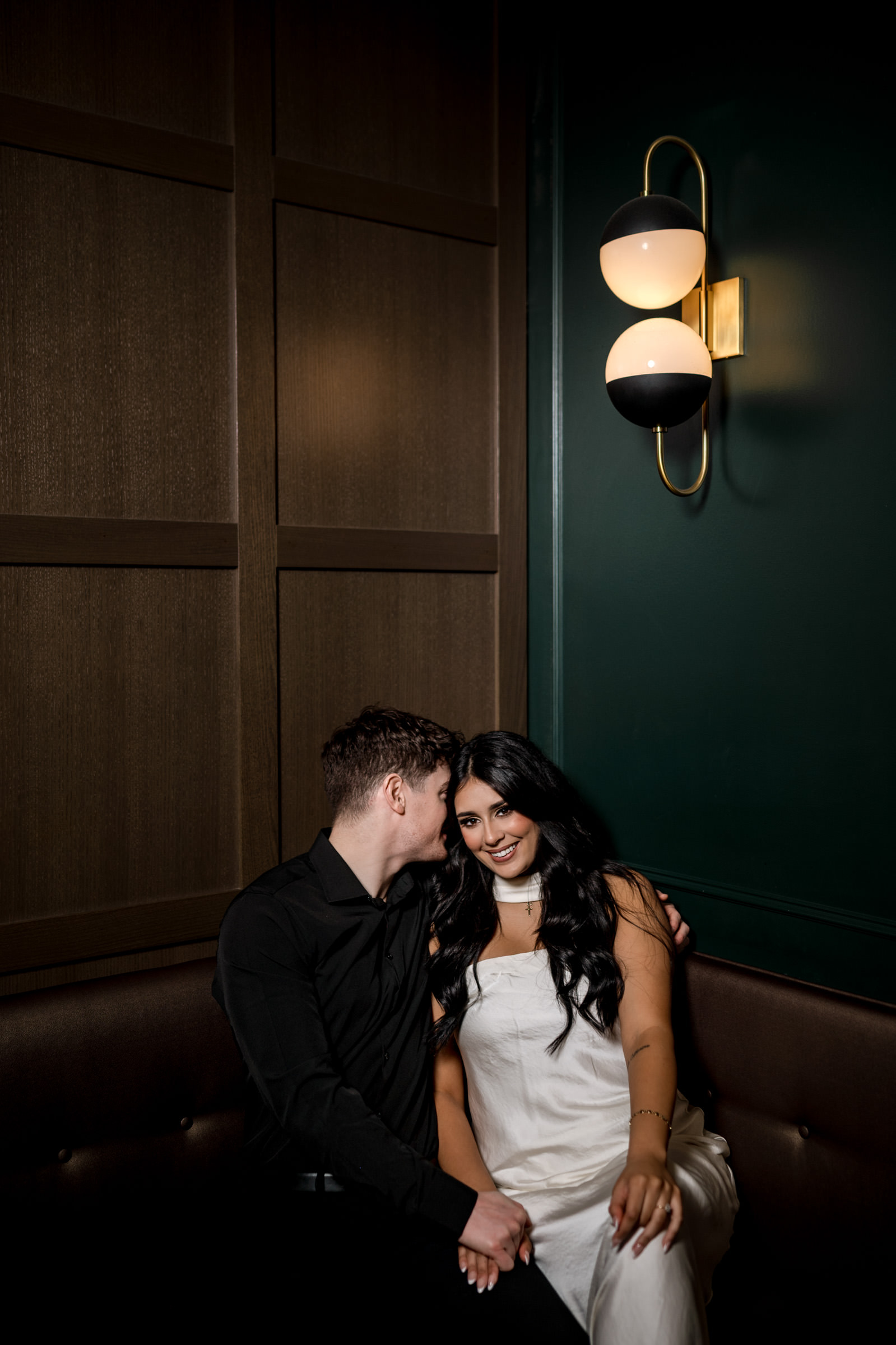 A couple enjoys their Winnipeg engagement on a bench in a dimly lit, wood-paneled room.