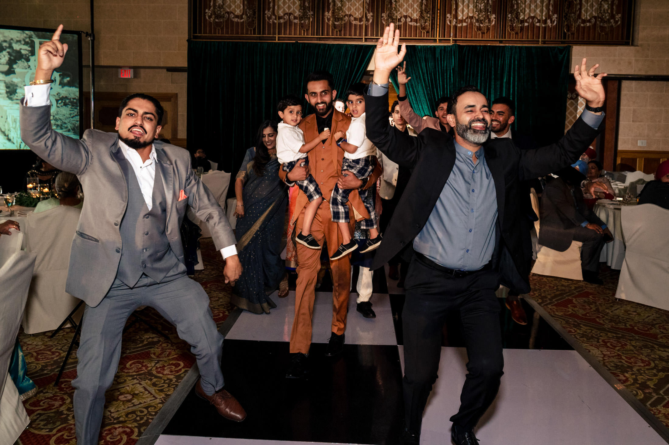 Men joyfully dance at a Winnipeg wedding; one holds two children.