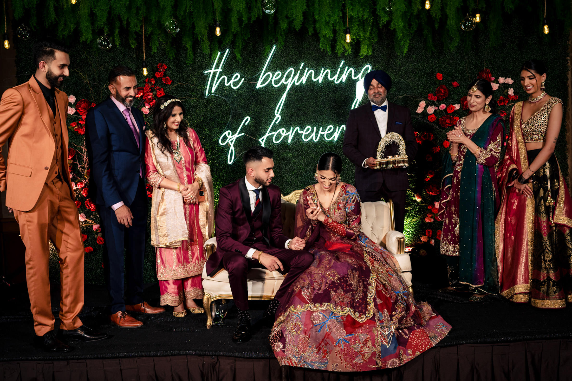 Bride and groom on stage with family at their Winnipeg wedding, under a neon "beginning of forever.