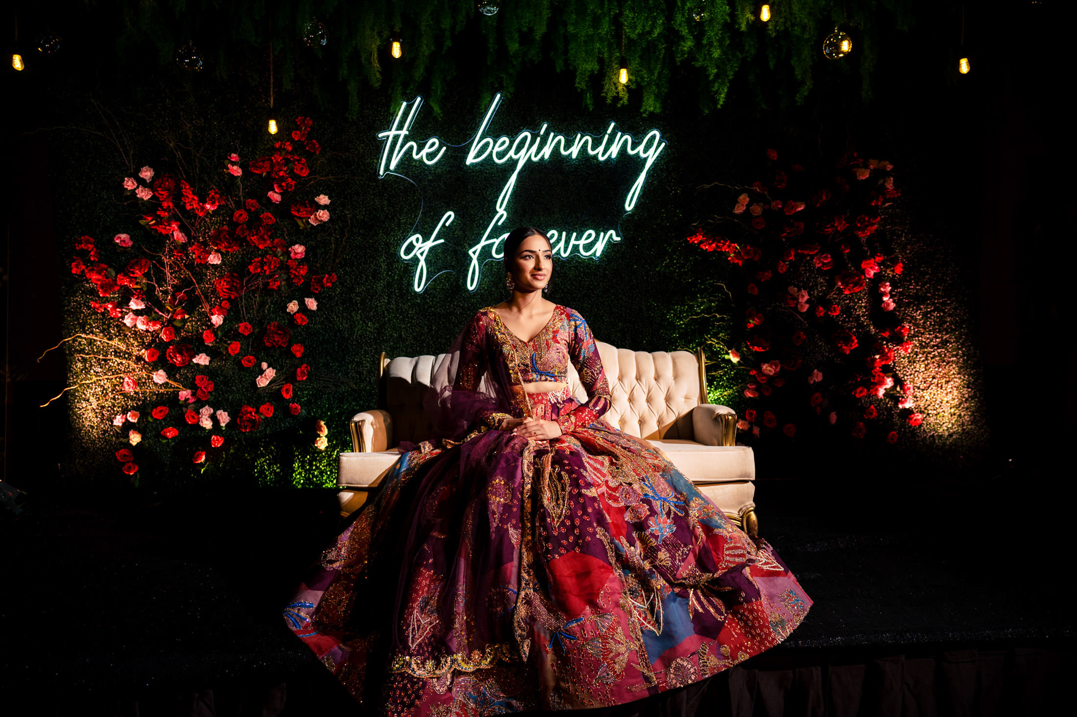Woman in colorful gown sits on sofa; neon "the beginning of forever" sign behind her at Winnipeg wedding.