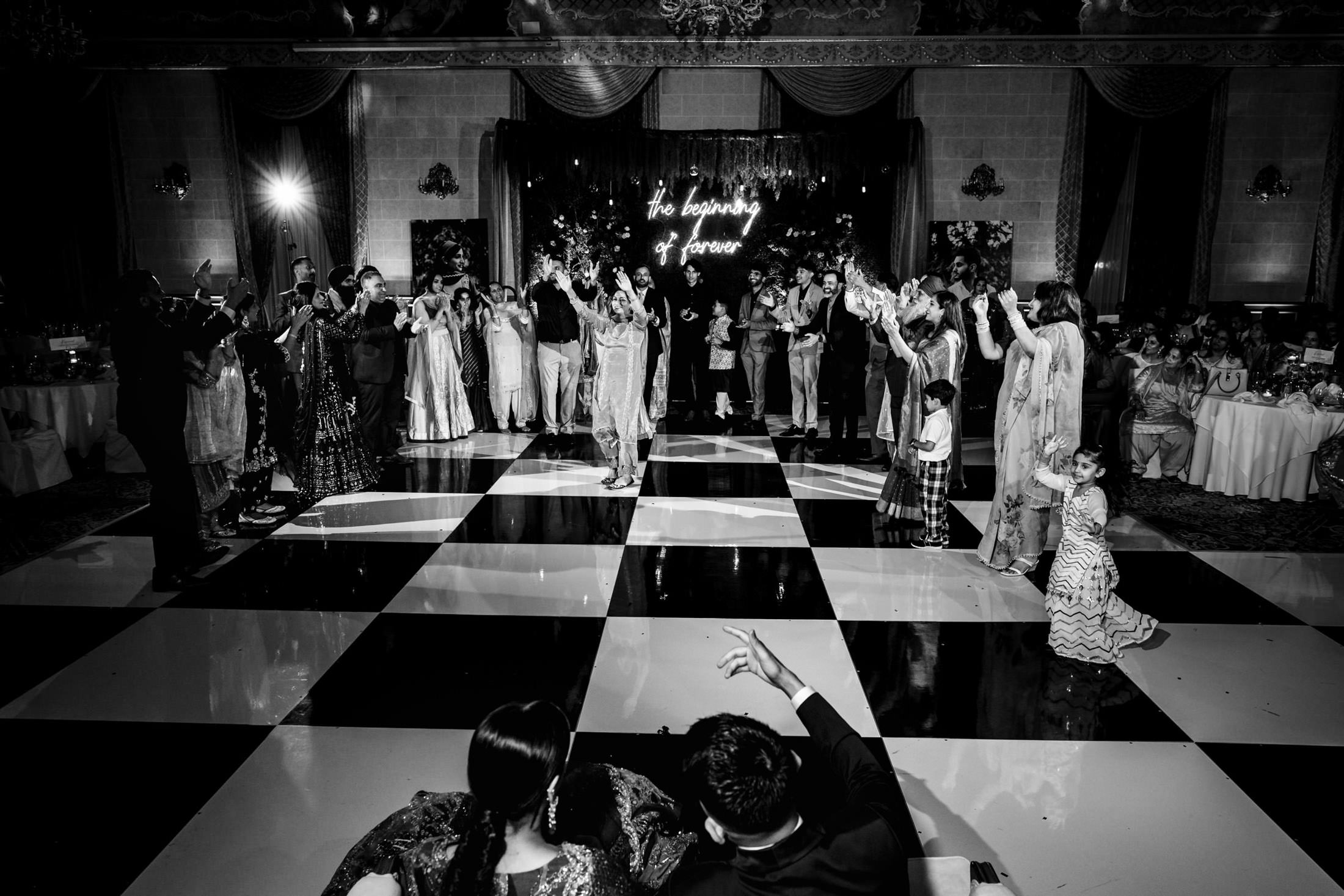 Guests dancing at a Winnipeg wedding on a checkered floor, beneath a glowing sign.