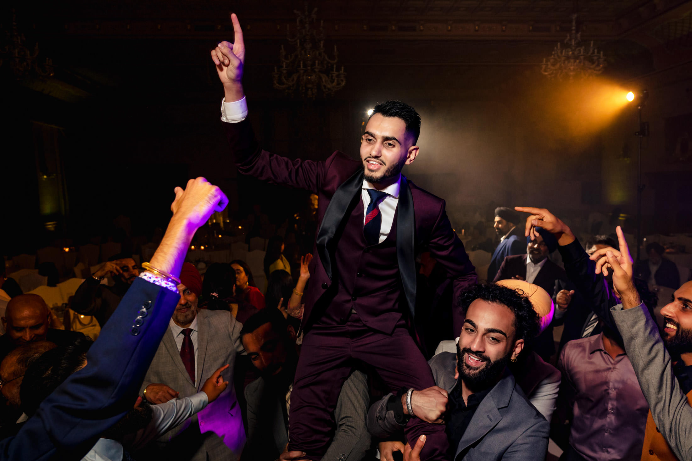 Person in a suit being lifted by a joyful crowd at a Winnipeg wedding with colorful lighting.