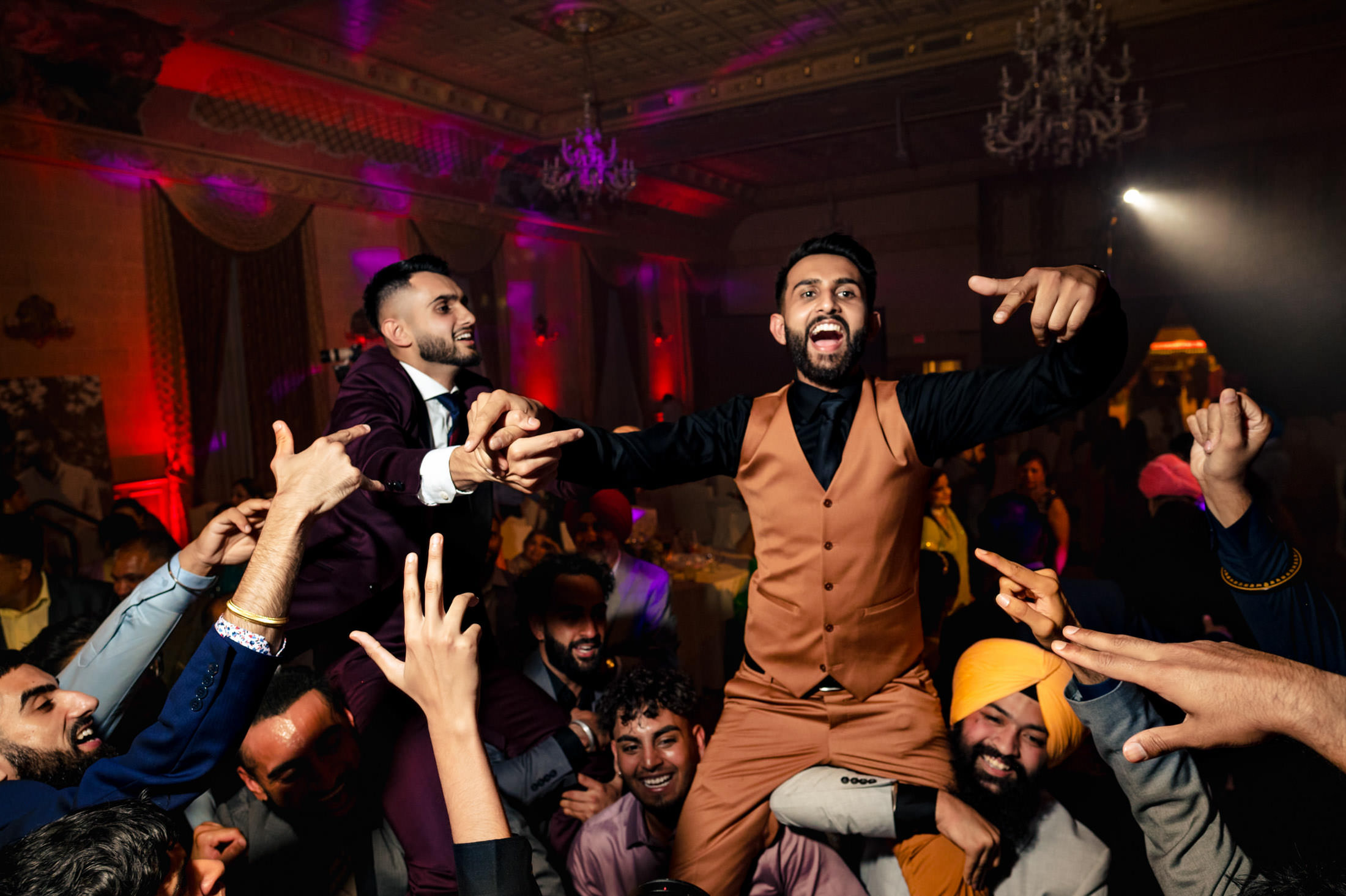 A joyful crowd lifts two smiling men on their shoulders at a lively Winnipeg wedding.