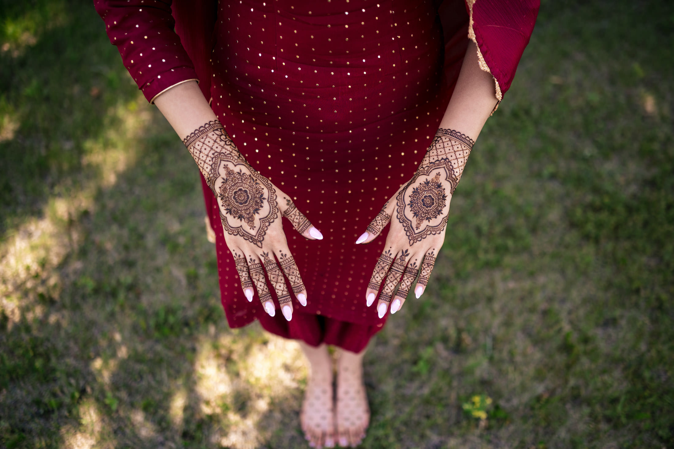 Hands with intricate henna designs, wearing a maroon dress, at a Winnipeg wedding on grass.