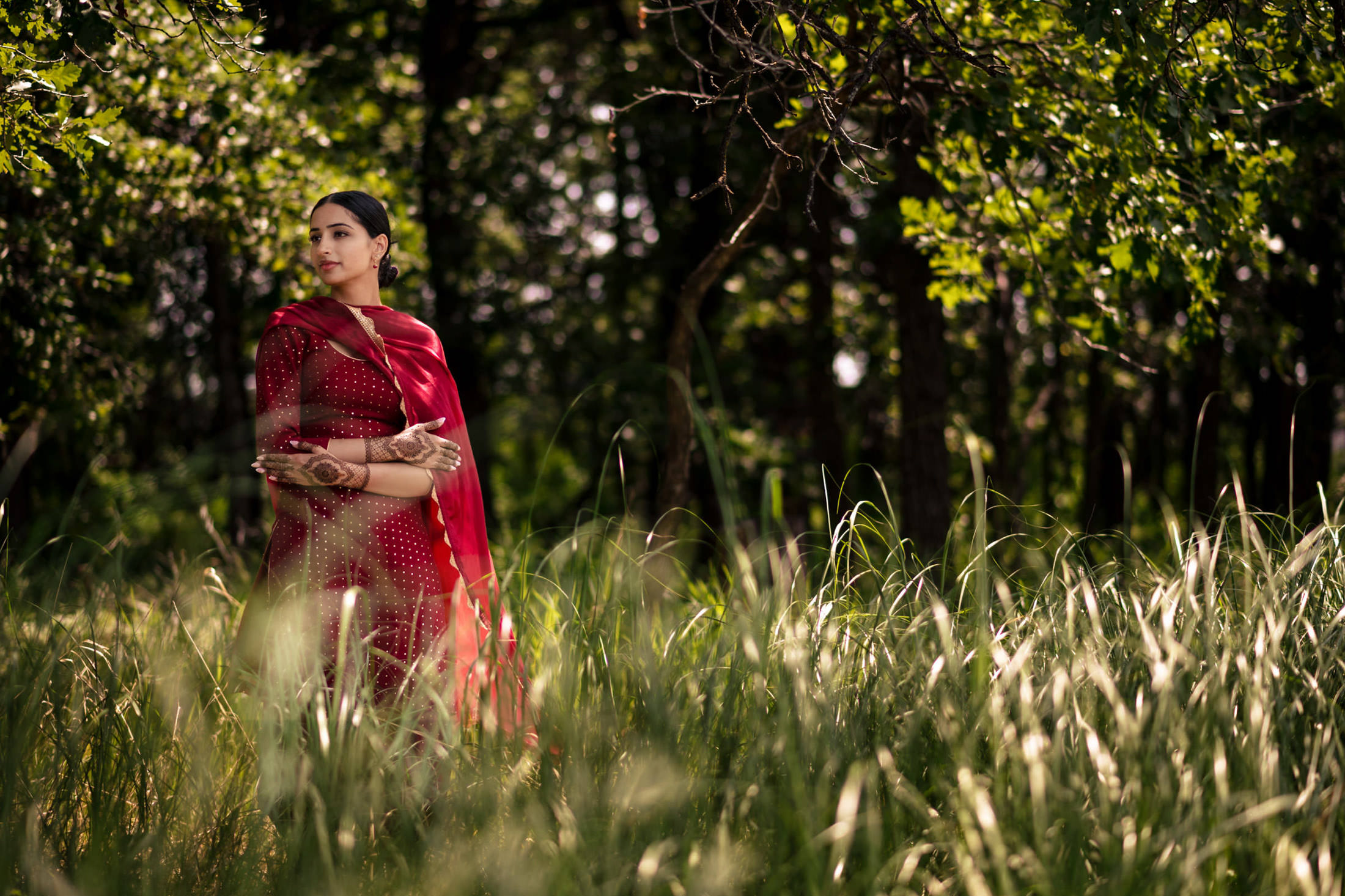 In a lush green forest, a woman in a red dress gazes aside; sunlight filters through.