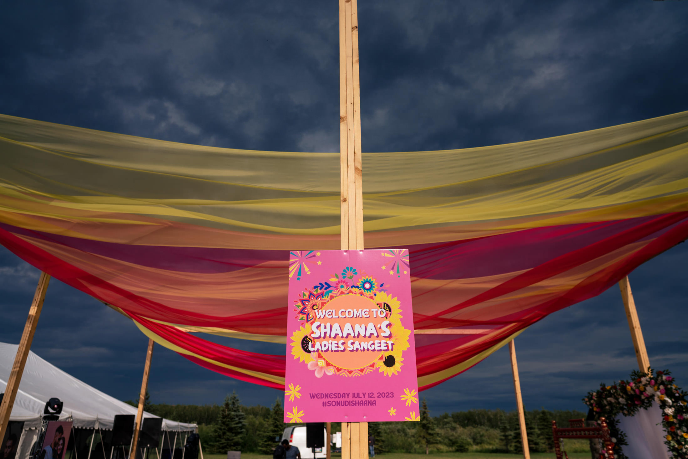 Colorful canopy with sign reading "Winnipeg Wedding: Shaana's Ladies Sangeet" under cloudy sky.