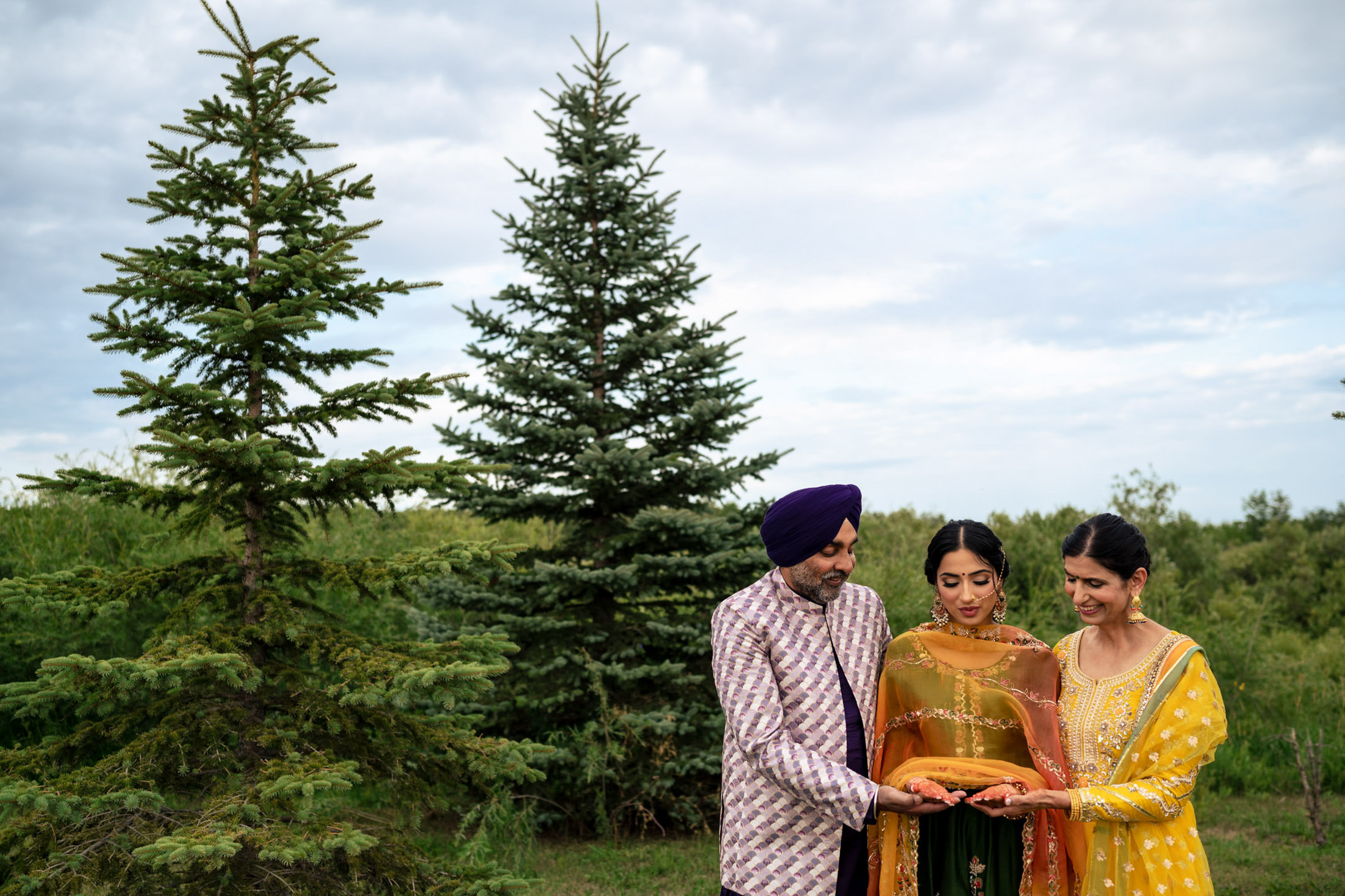 Three in traditional attire join hands near trees under a cloudy Winnipeg wedding sky.