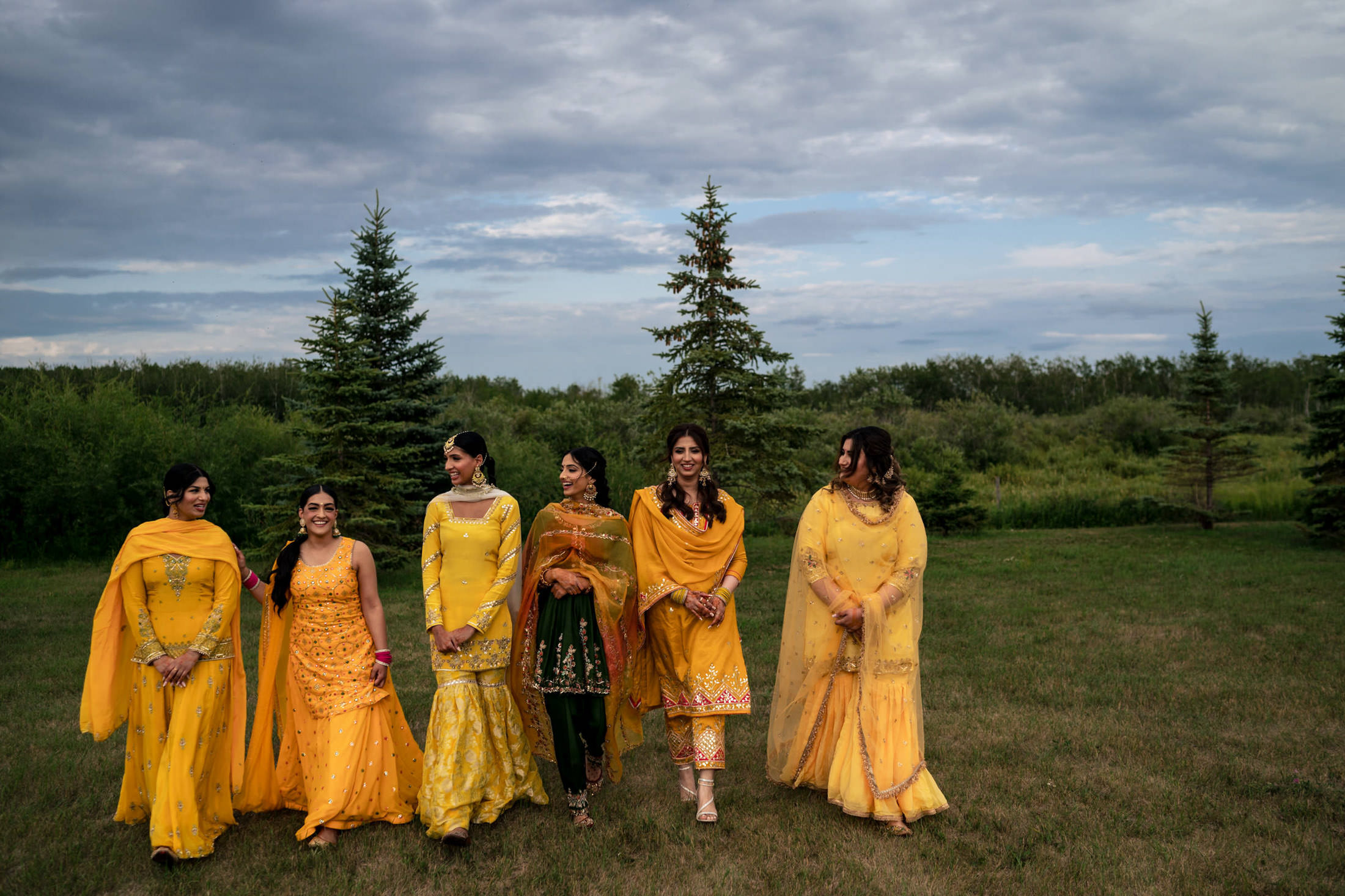 Six women in yellow outfits stroll on grass under Winnipeg’s cloudy wedding sky.