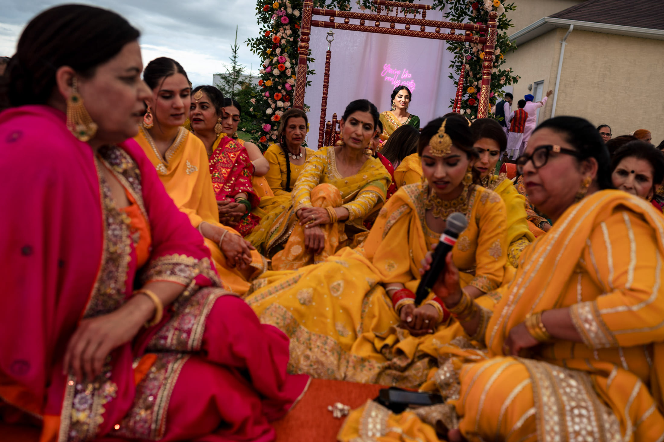 Women in colorful dresses at a Winnipeg wedding, one holding a microphone, outdoors.