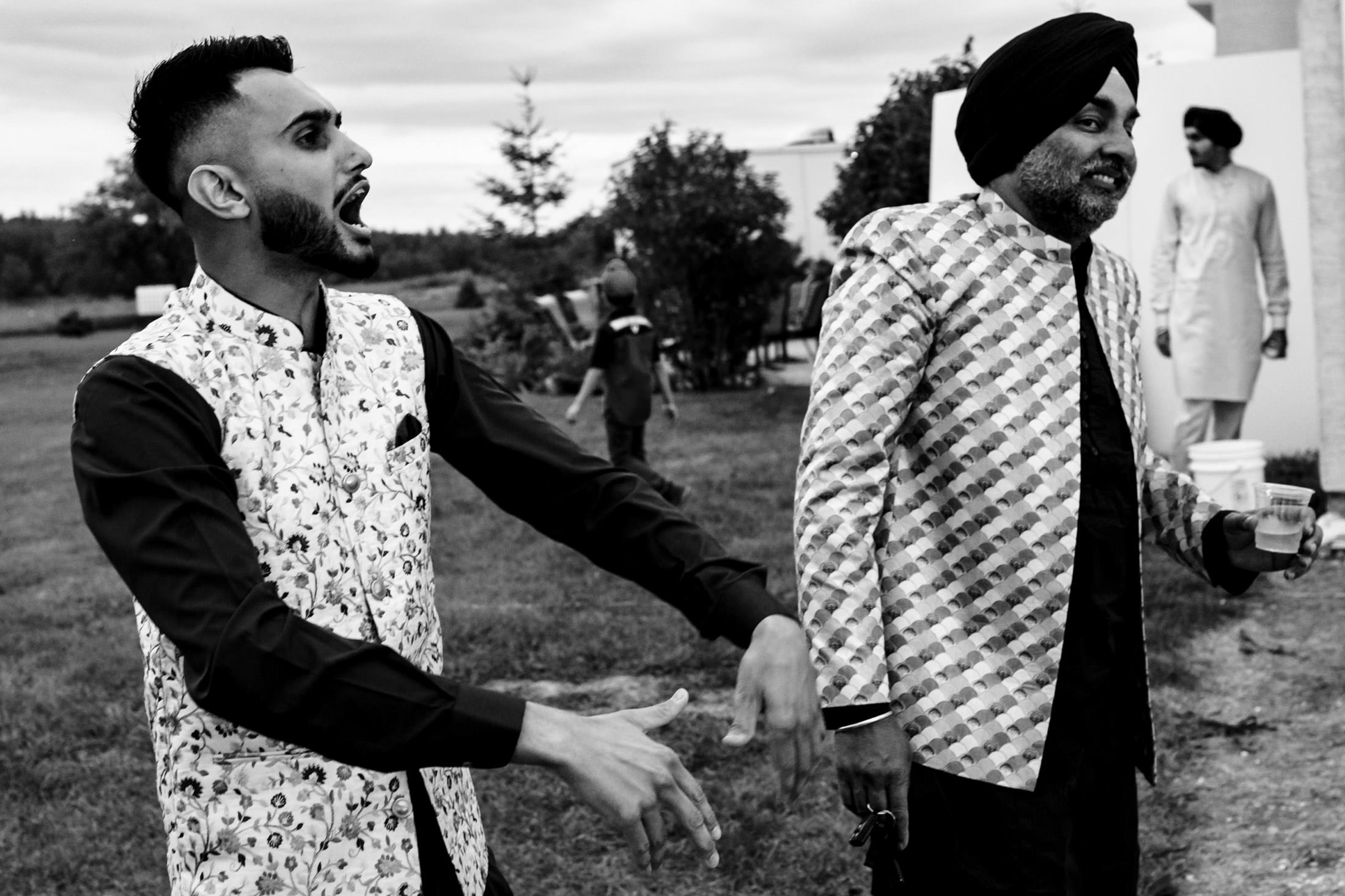Two men outdoors in festive attire at a Winnipeg wedding, one speaking energetically, the other smiling.