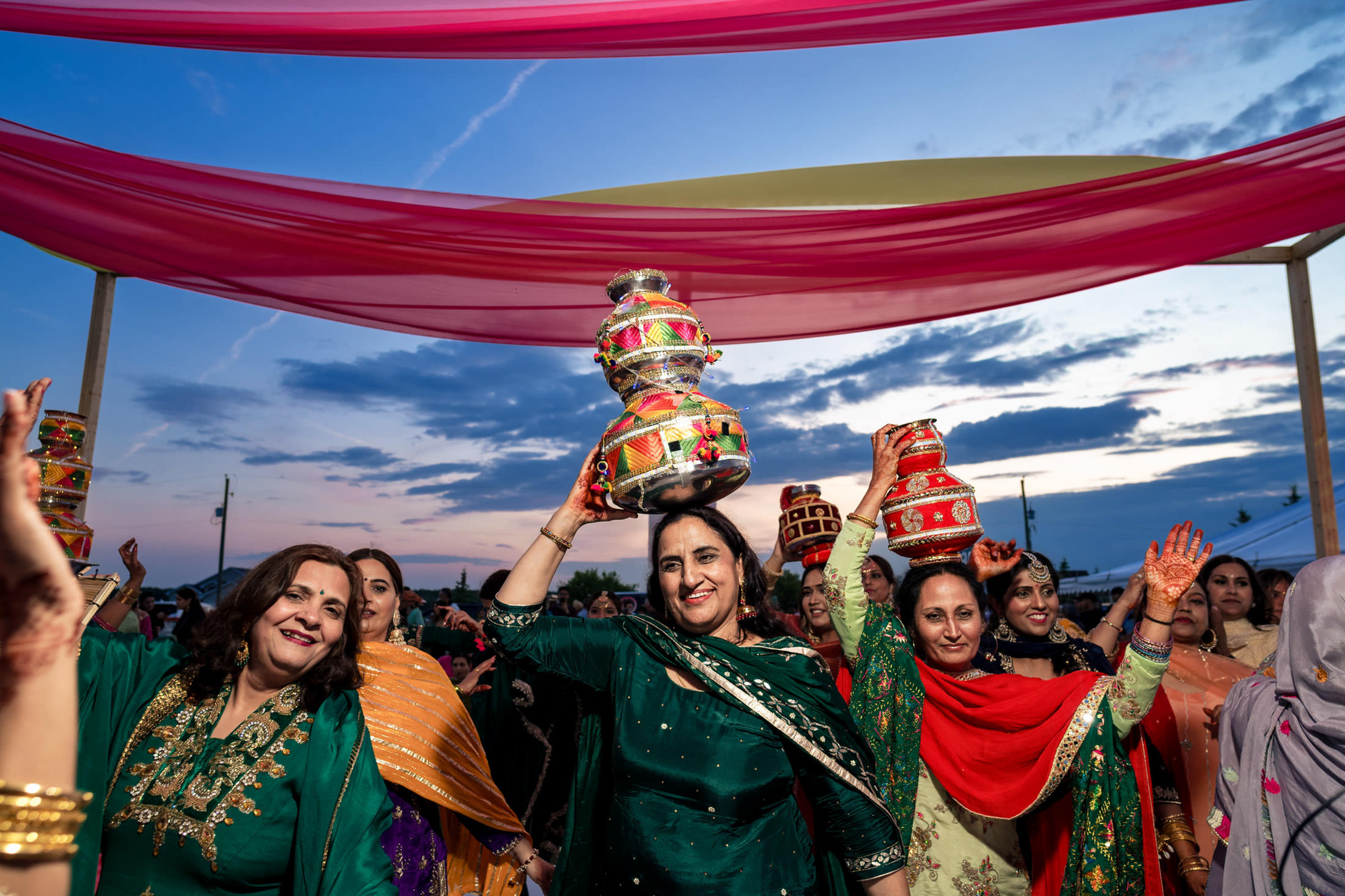 Women in festive attire at a Winnipeg wedding, carrying pots under colorful fabric canopies.