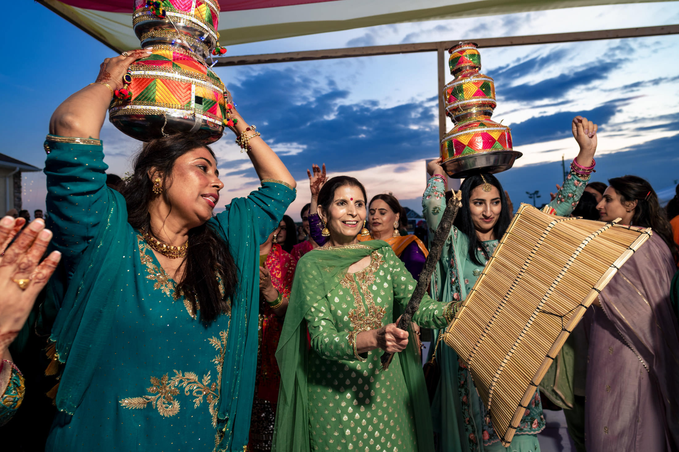 Women in colorful dresses dance with pots and a fan under a vibrant Winnipeg wedding sky.