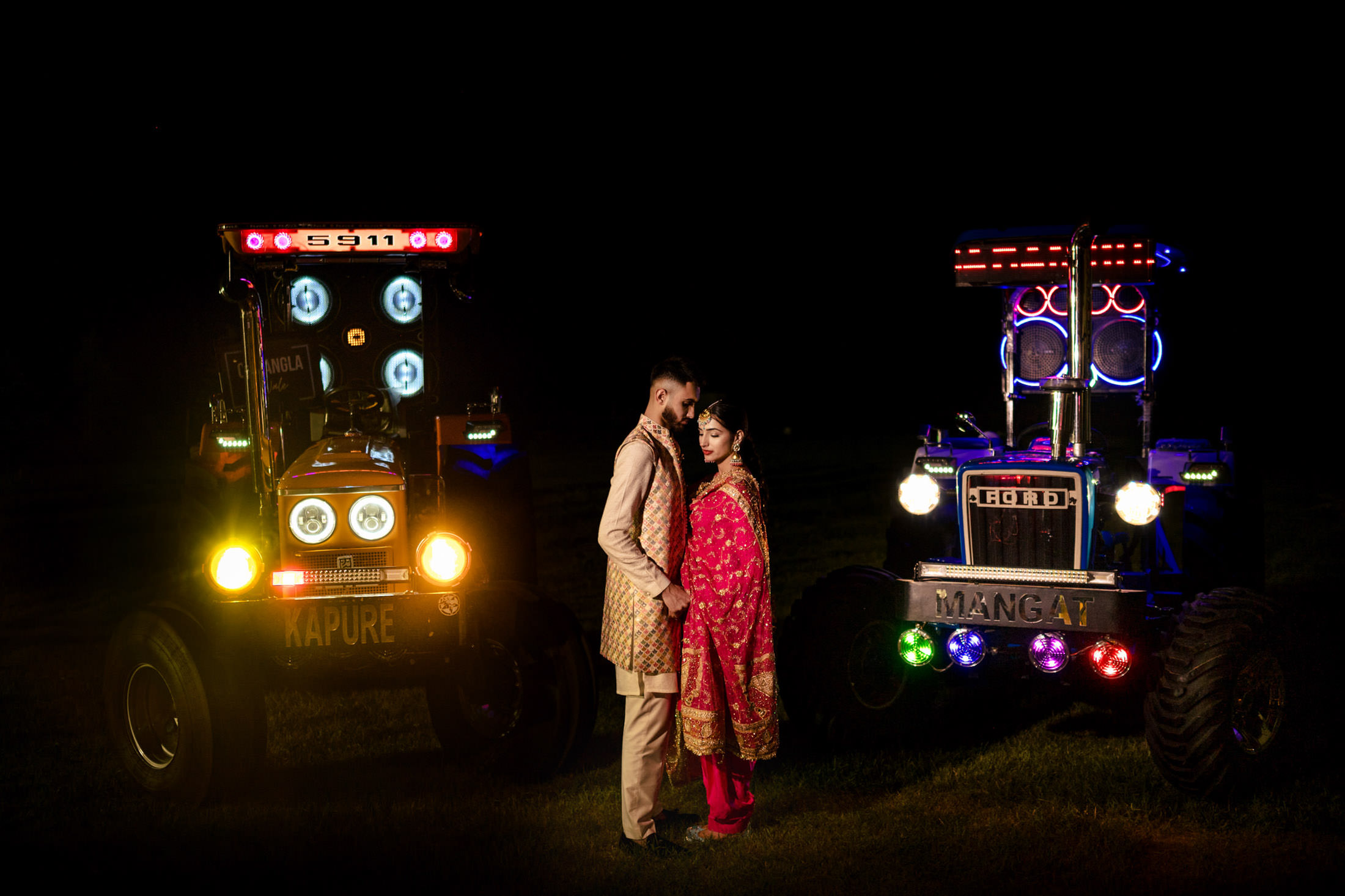 Couple in traditional attire pose between two illuminated tractors on a Winnipeg wedding night.