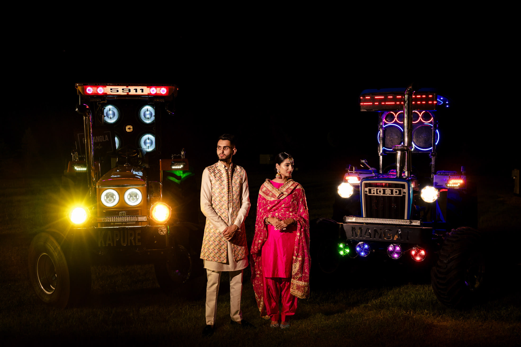 A Winnipeg wedding couple in traditional attire stands between two illuminated tractors at night.