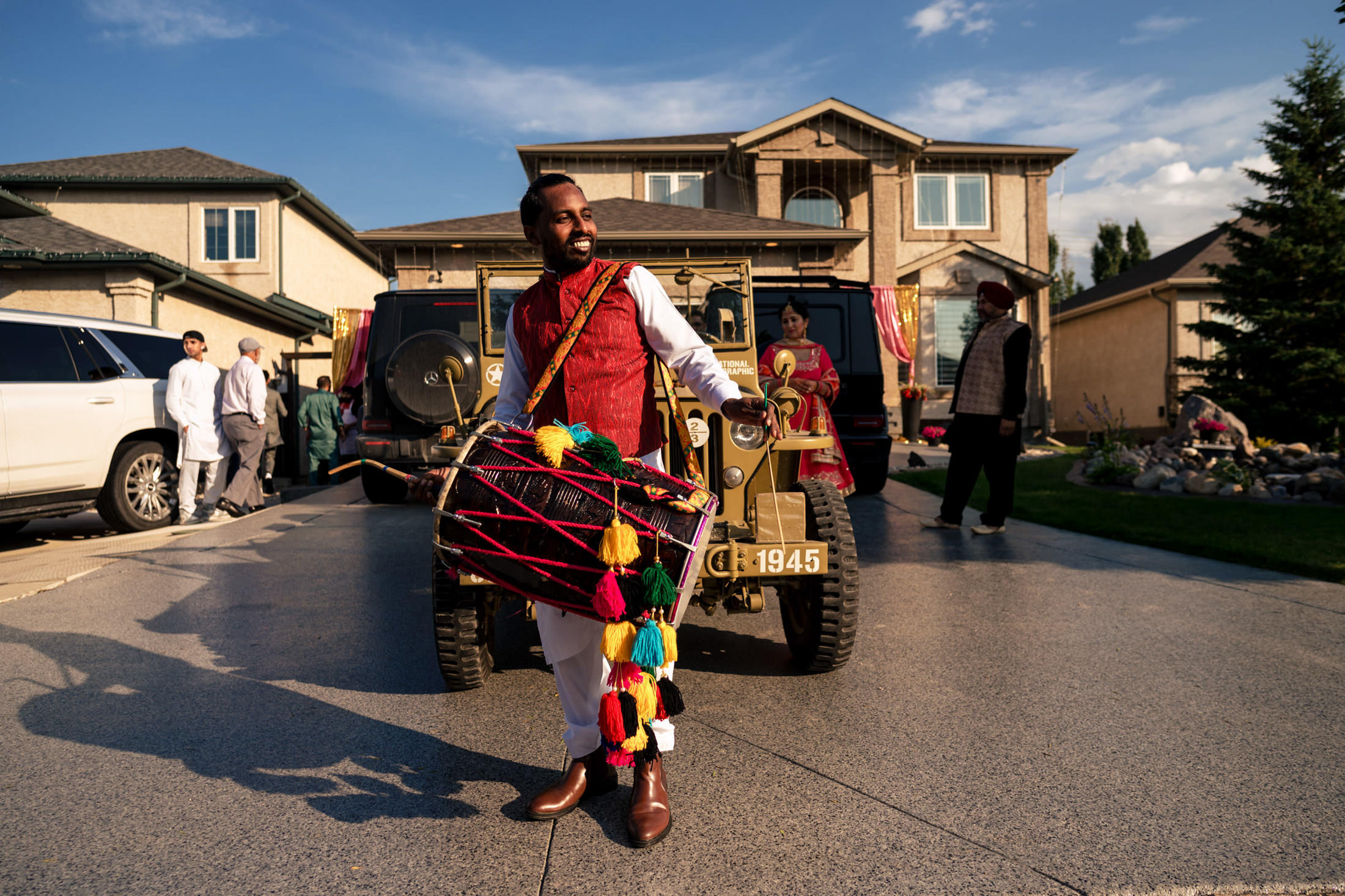 Man playing colorful dhol drum at a vibrant Winnipeg wedding, surrounded by people and cars.