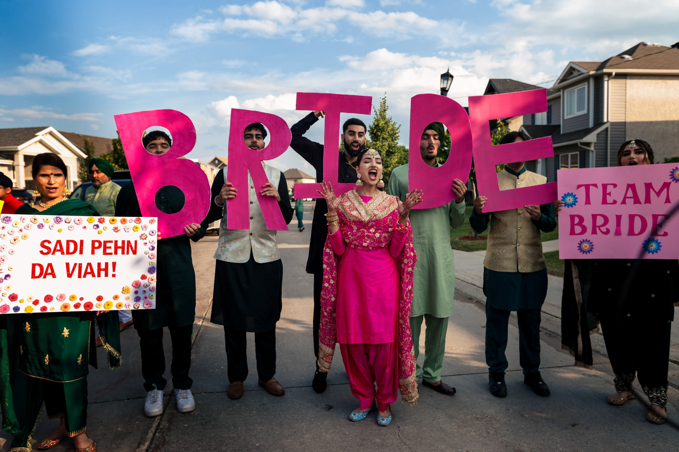 Group in Winnipeg holding "BRIDE" and "Team Bride" signs in colorful traditional attire.