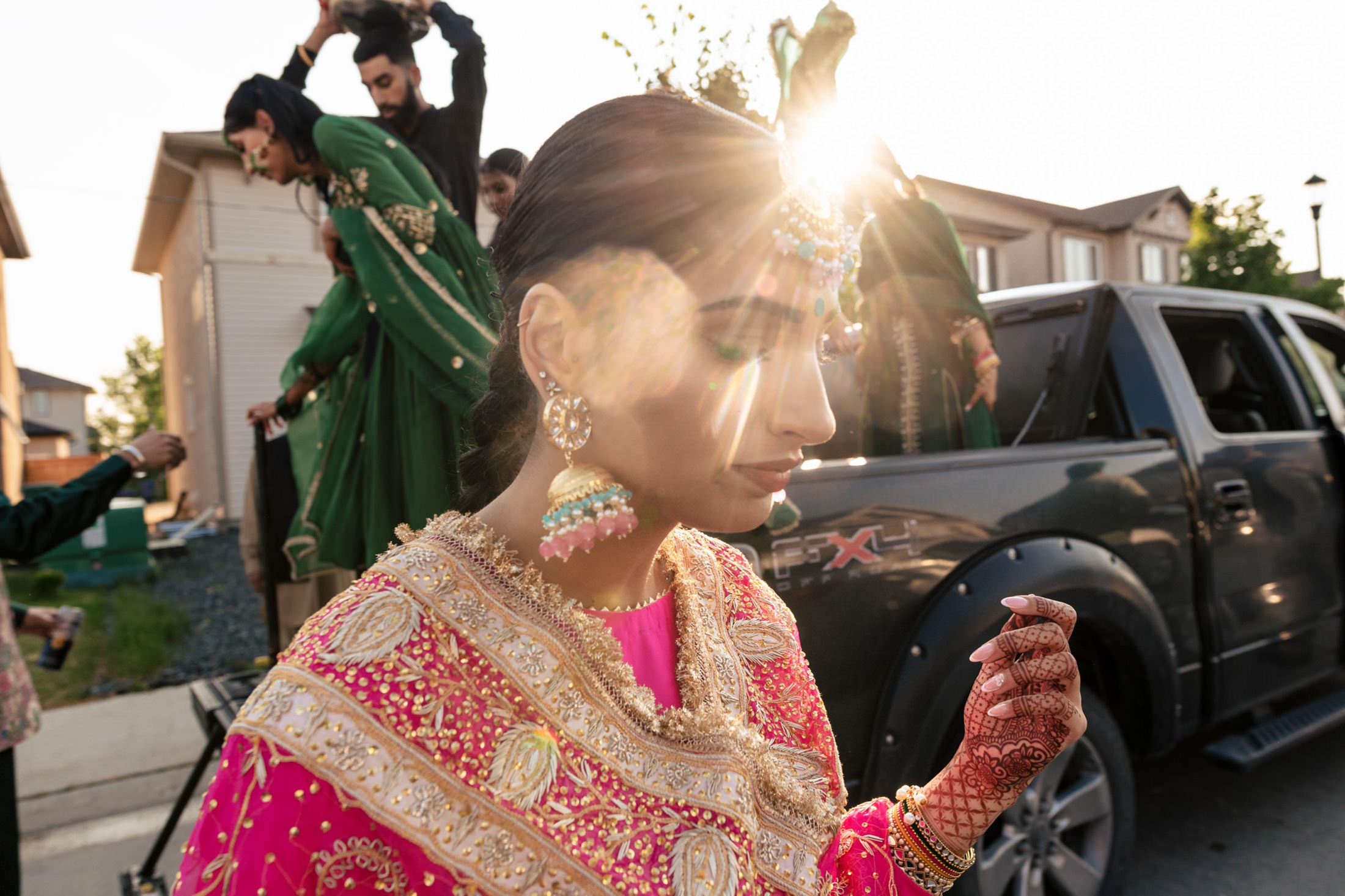 Person in ornate attire with sunlight flare; others in green outfits, like a Winnipeg wedding.