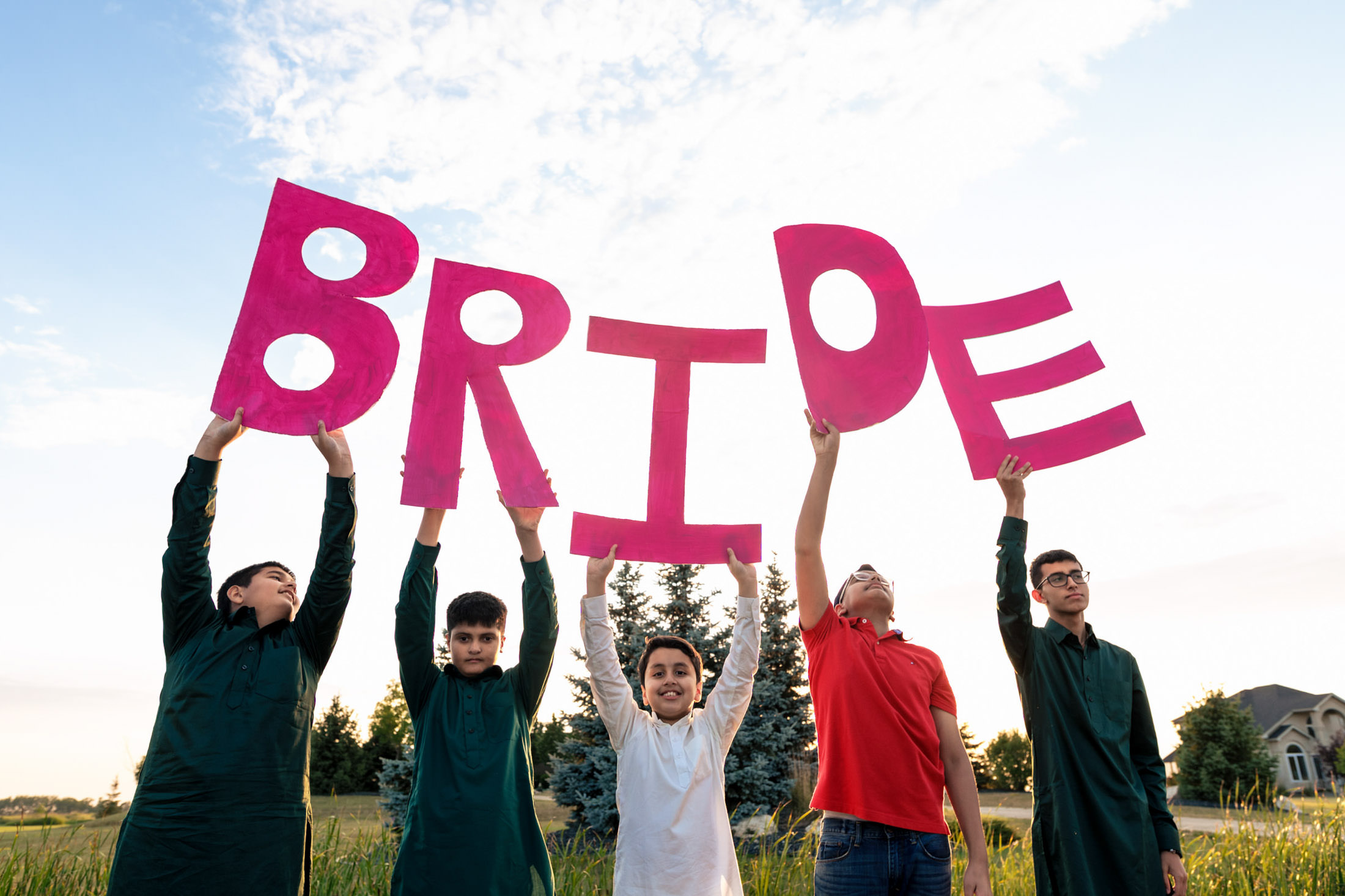Five people outdoors hold pink letters spelling "BRIDE" under Winnipeg's cloudy sky.