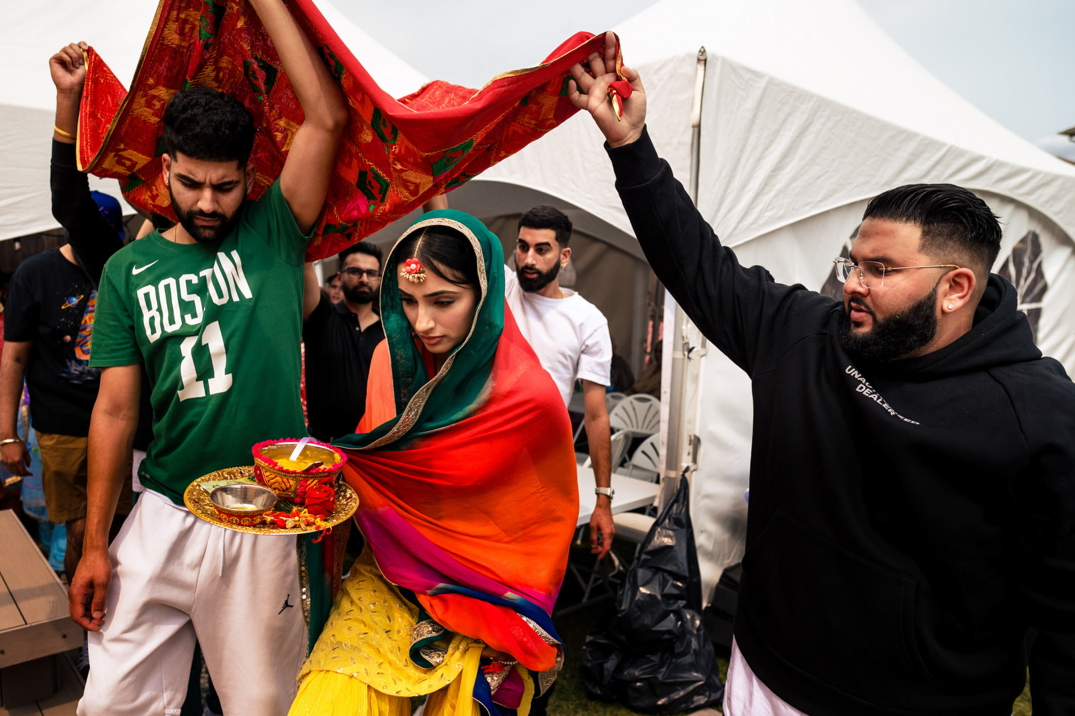 A woman in colorful attire under a red cloth, flanked by two men, at a Winnipeg wedding ritual.