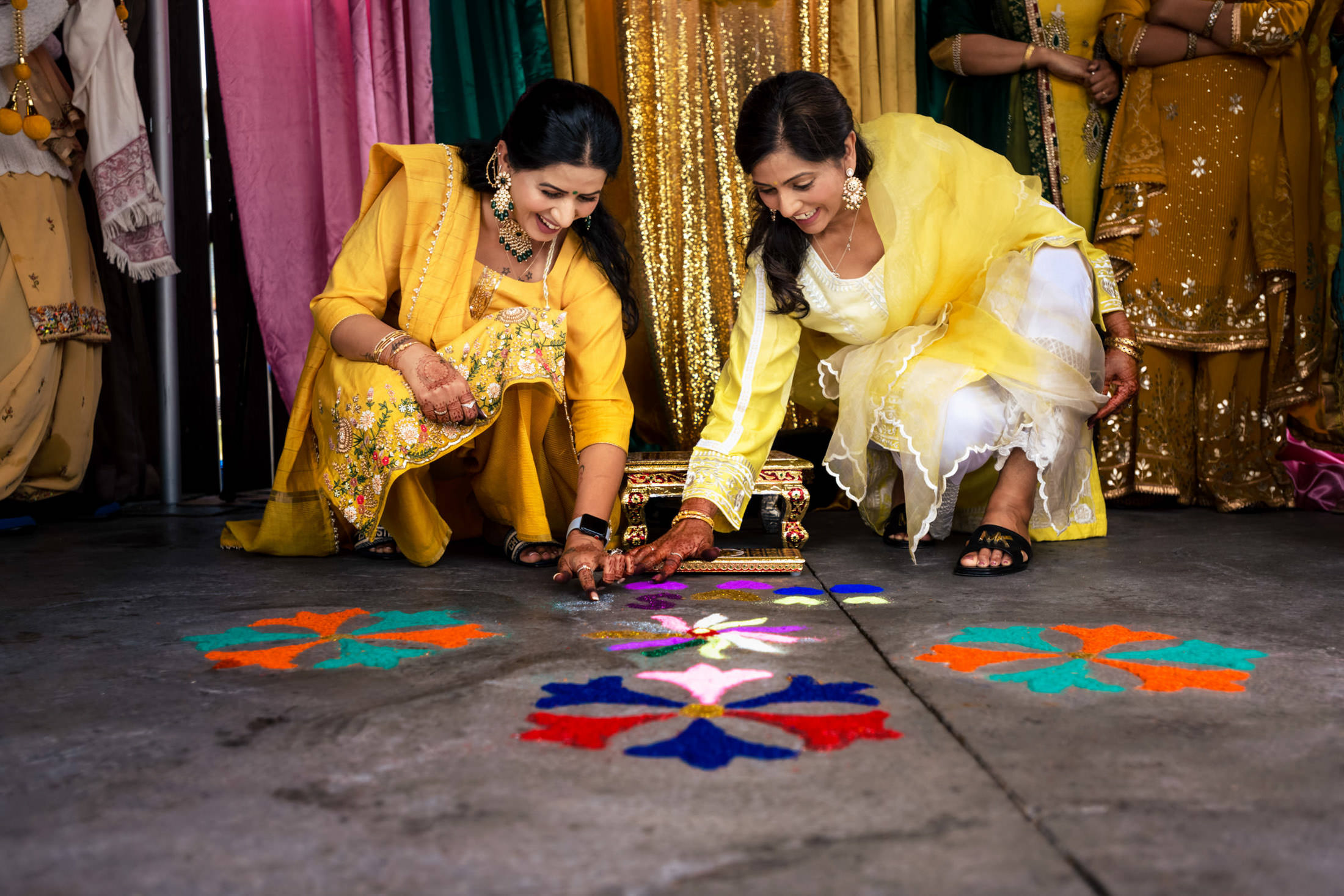 Two women in colorful attire craft a vibrant floor design with powders at a Winnipeg wedding.