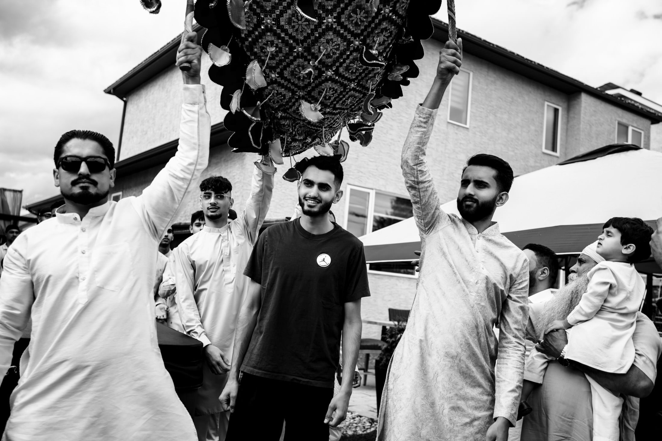 Men celebrate outdoors under a decorated canopy at a Winnipeg wedding, child on shoulder.
